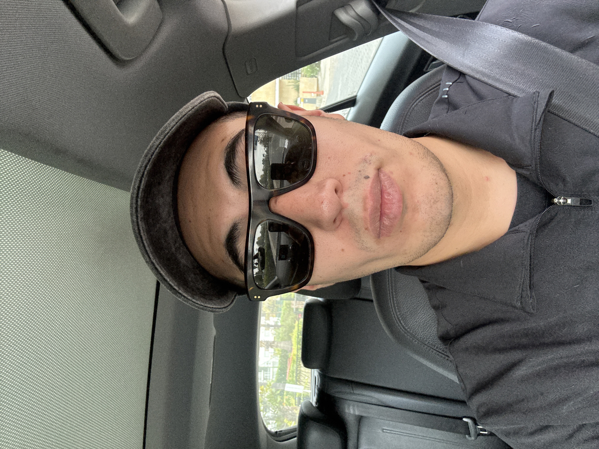 A self-portrait captured inside a car shows a person, likely male, from the chest up. The image is oriented with the person's head tilted to the left, as if lying on their side. The individual wears dark sunglasses with tortoise-shell frames, a dark grey or black cap, and a dark grey or black jacket with a zippered collar. A seatbelt is visible across their chest, secured into a buckle.
The person's face shows a neutral expression, with their lips slightly parted. Stubble is visible on their chin and jawline, and there are a few small moles or freckles on their face. The sunglasses reflect the interior of the car and some indistinct outdoor scenery.
The setting is inside a vehicle, indicated by the car's headliner, a grab handle, and part of a car seat or headrest. Through the side windows, blurred outdoor scenery can be seen, including green foliage and parts of light-colored buildings or structures, typical of an urban or suburban environment. The light suggests it is daytime, possibly under an overcast sky or with some diffusion from window tinting or a sunroof screen, creating a soft, even illumination. Given the location context, this scene is set in San Bernardino Tlaxcalancingo, Mexico. No visible text is present in the image.