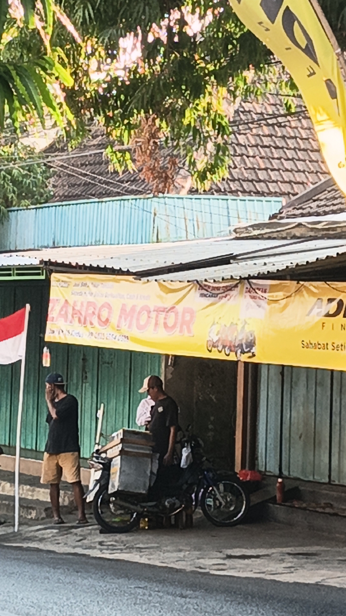 The image depicts an outdoor scene in Dawe, Indonesia. A yellow banner with red lettering that reads "ZAHRO MOTOR" is prominently displayed above a shop. To the left of the banner, a red and white Indonesian flag is visible. Two men are standing near a motorcycle parked on the side of a road. One man, wearing a black t-shirt and khaki shorts, stands with his hand covering his face. The other man, in a dark t-shirt and baseball cap, stands next to the motorcycle which has a cargo box attached to its rear. The background shows a corrugated metal roof and green corrugated metal siding. The overall setting appears to be a commercial area on a sunny day.