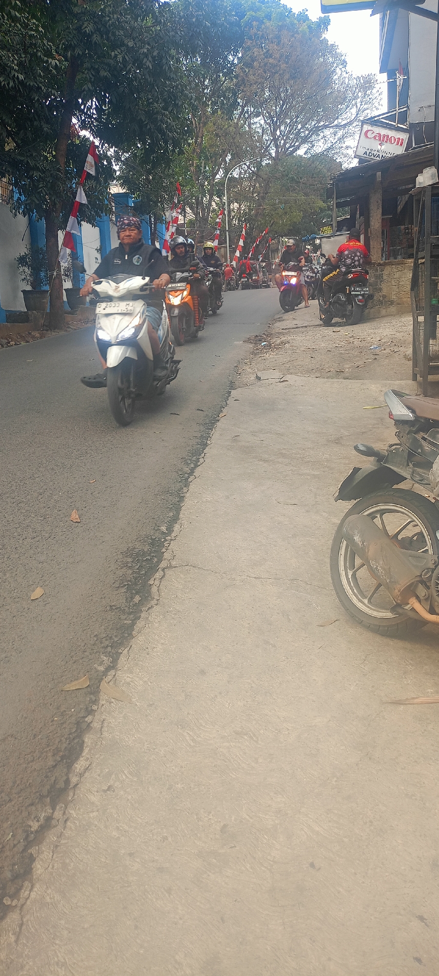 The image captures a street scene in Bandung City, Indonesia, likely during an event or parade given the presence of numerous motorcycles and Indonesian flags. In the foreground, a man rides a white scooter, wearing a dark shirt and a head covering. He is followed by several other individuals on motorcycles, some wearing helmets and casual clothing. The motorcycles are traveling in a line along a paved road that transitions into a concrete sidewalk. Trees line the left side of the road, with a blue and white building visible behind them. Red and white Indonesian flags are prominently displayed on poles along the street. In the background, a building with a "Canon" sign is visible, suggesting a commercial area. The overall atmosphere appears to be one of movement and gathering, possibly a procession or a group ride.