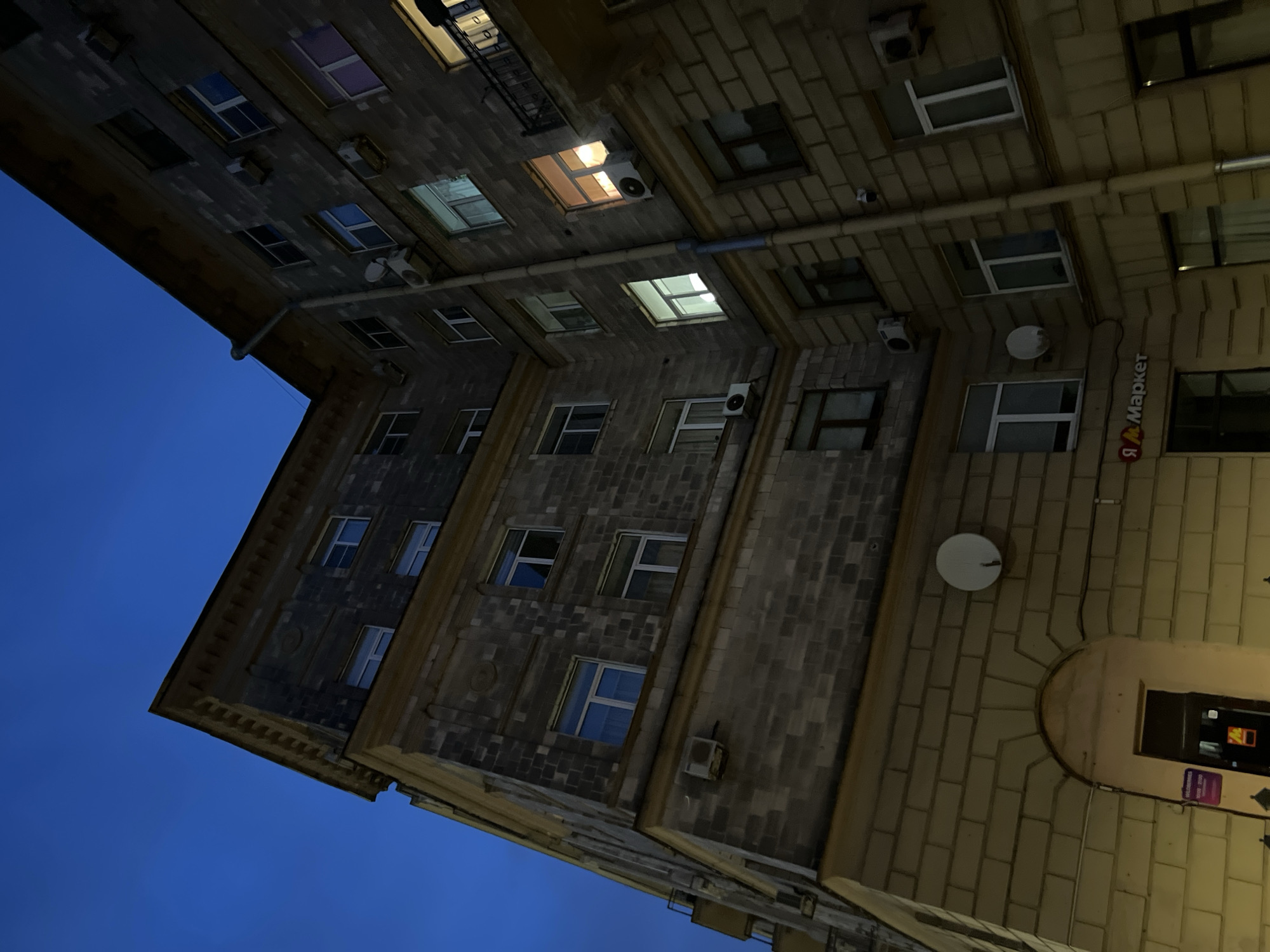 The image shows a night view of an apartment building in Saint Petersburg, Russia, taken from a low angle looking up. The building appears to be made of light-colored stone or concrete blocks, with some darker, textured stone on the upper floors. The sky is a deep, dark blue, indicating it is either very early morning or late evening.
The building has multiple floors with many windows. Some windows are dark, while others are illuminated, suggesting inhabitants are present. Several windows have air conditioning units installed below them. There are also what appear to be satellite dishes mounted on the exterior walls.
On the lower level of the building, there is an arched entrance with a door. Above this entrance, a sign reads "Я Маркет" (Ya Market), indicating a retail establishment. A purple sign with Cyrillic text is also visible near the entrance. The lighting at street level is warm and yellow, illuminating the lower facade of the building.
The architectural style suggests an older building, characteristic of Saint Petersburg. The overall scene is typical of urban residential areas at dusk or dawn.
