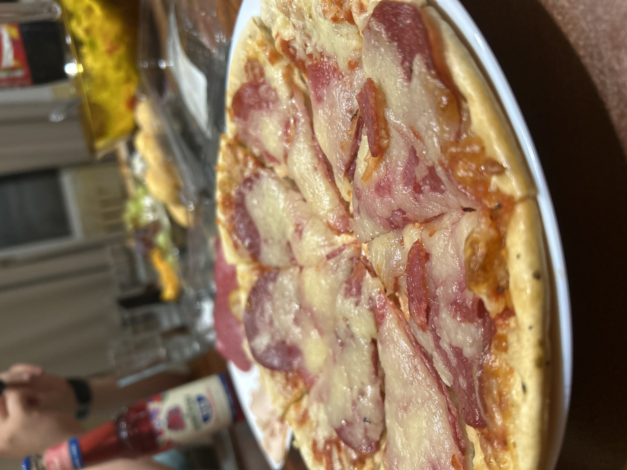 The image displays a close-up, slightly angled view of a round pizza, sliced and presented on a white plate. The pizza is generously topped with melted white cheese and distinct, reddish-pink slices of cured meat, resembling salami or pepperoni. The crust is golden brown and appears to be seasoned.
In the foreground, a person's hand and wrist, adorned with a dark band, hold a red bottle. The bottle features a white and blue label, clearly showing the Cyrillic word "Джем" (Jam) and part of the brand name "Марица."
The background is softly blurred but reveals an indoor setting, consistent with a home or casual dining area. Hints of other items, including what appears to be a snack or pastry in a yellow wrapper, are visible on a surface behind the pizza. The lighting suggests it is evening or nighttime, relying on artificial light sources.
The scene depicts a casual meal or snack, with someone either about to eat the pizza or in the process of dining. The presence of the bottle suggests the person might be adding sauce or a condiment to their food or having a drink alongside. The overall atmosphere is relaxed. While no specific visual details within the image point to the location, the provided context places the scene in Sofia, Bulgaria. Faintly visible on a background surface are the numbers "99".