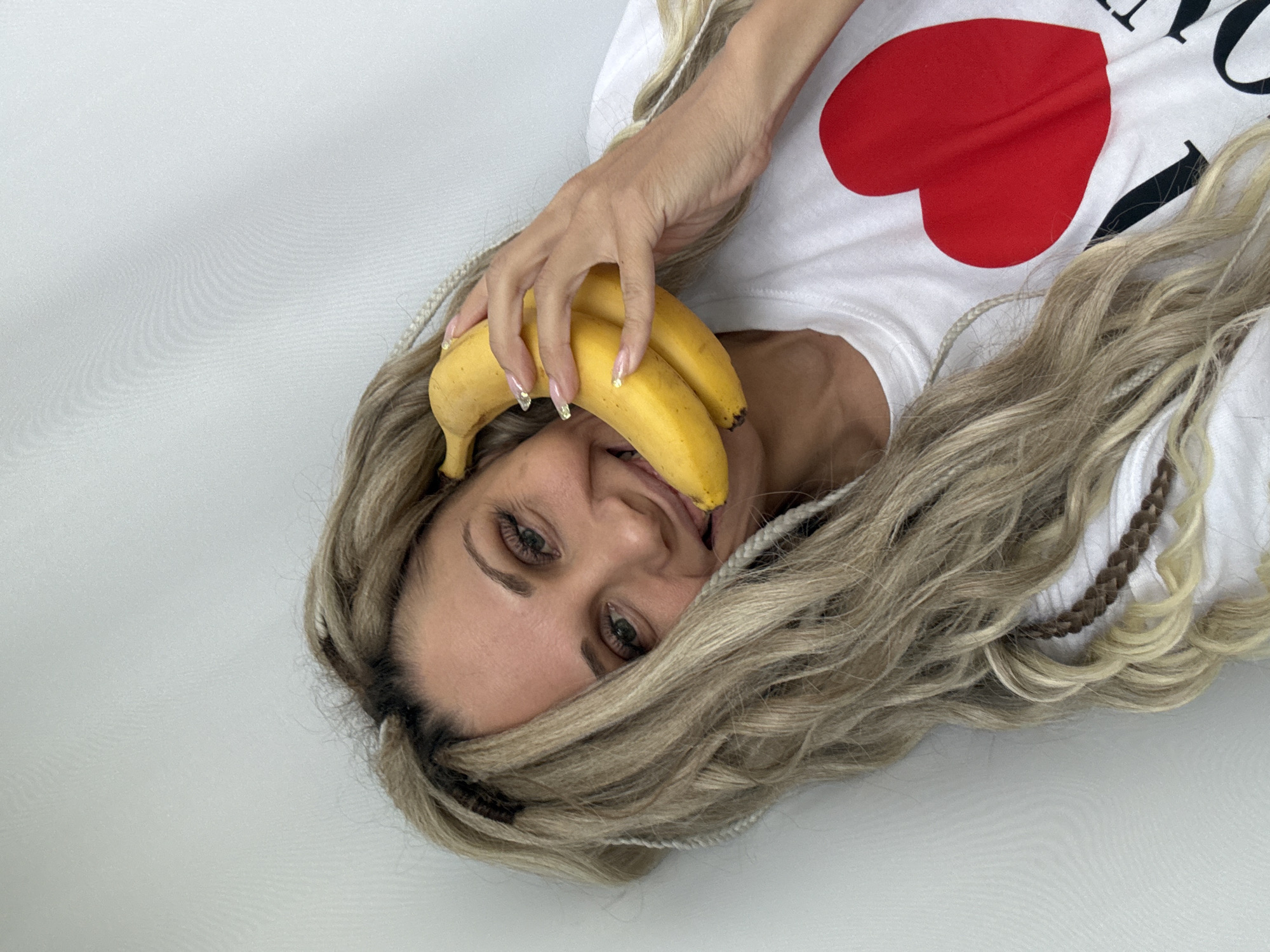 A light-skinned woman with braided blonde hair is holding three bananas in her right hand, with one of the bananas held in her mouth. She is wearing a white t-shirt with a red heart design. The image background is white. No location information for Rusanovo, Russia, is determinable from the provided image.