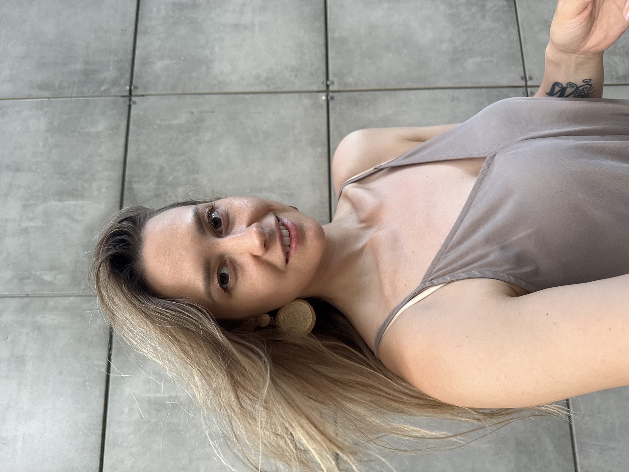In the city of Nebug, Russia, a medium-shot photograph shows a blonde woman with her hair blowing in the wind, standing in front of a grey, tiled wall. She is wearing a light brown, spaghetti-strap dress and large, circular, woven earrings. The woman is smiling and looking towards the camera. A tattoo is visible on her left wrist.
