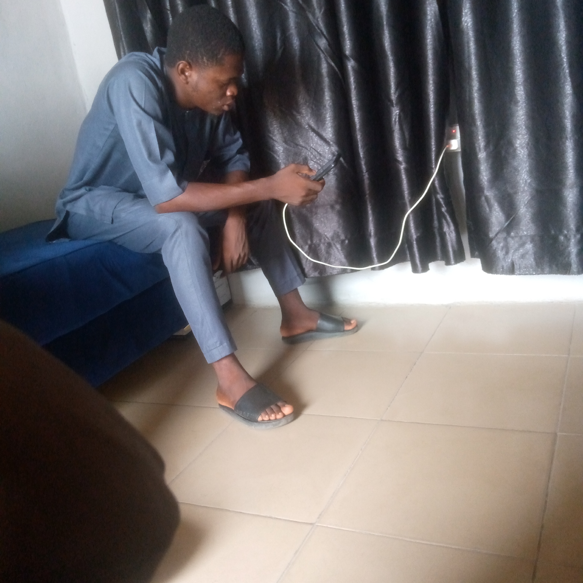 A young Black man in a blue tunic and trousers is sitting on a blue upholstered bench. He is holding a smartphone in his right hand and looking at it. The phone is plugged into a white power adapter which is inserted into a white electrical outlet on a dark grey curtain. The curtain is hanging behind the man, and it appears to be pleated. The man is wearing black slide sandals. He is sitting in a room with light-colored tiled flooring. The lighting appears to be artificial.