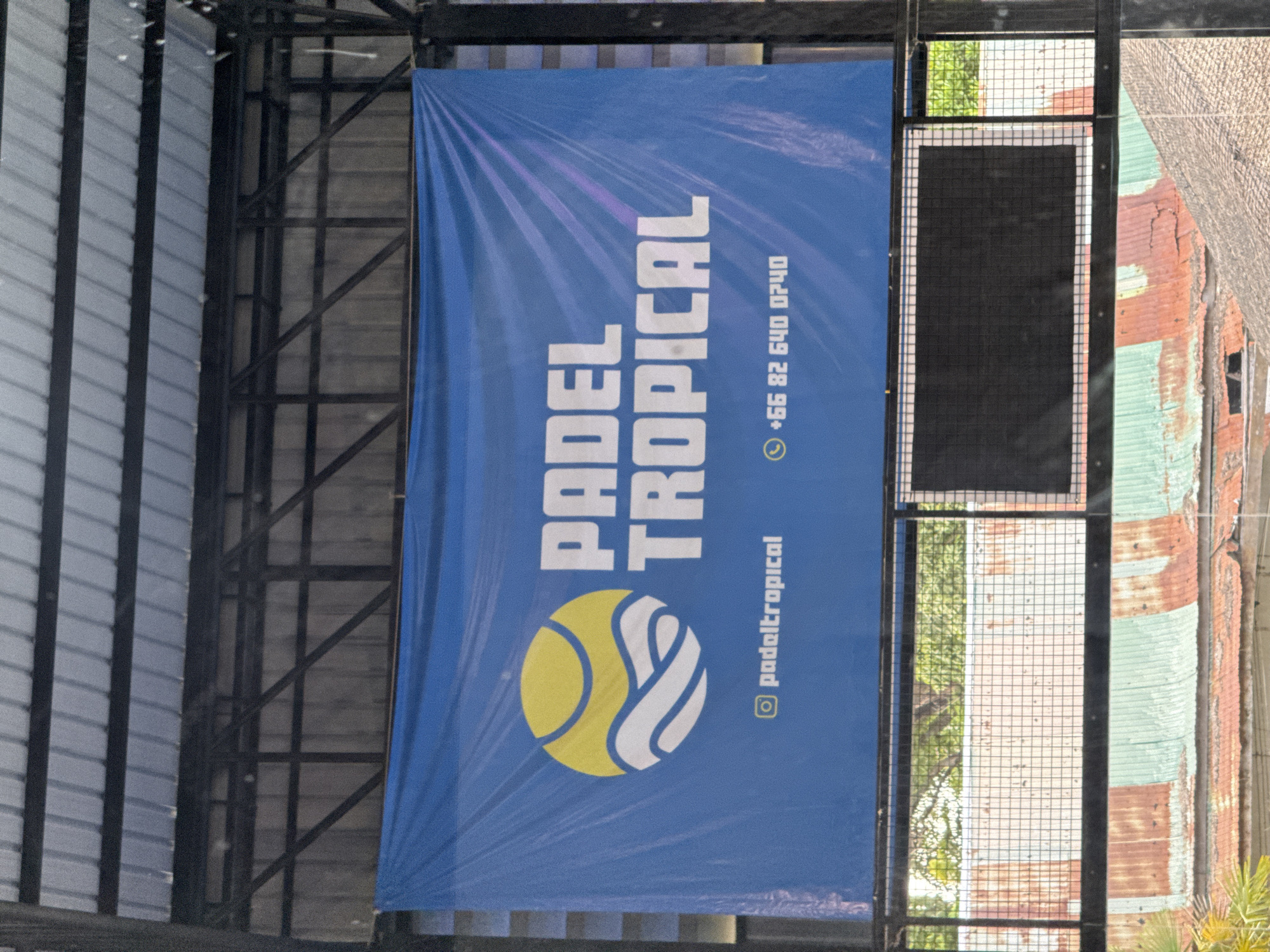 A blue banner hangs from the metal rafters of an outdoor or semi-outdoor structure. The banner displays the logo and name of "PADEL TROPICAL" in white text. To the left of the text is a circular logo with a yellow sun-like orb and white wavy lines. Below the logo and text, the Instagram handle "@padeltropical" and a phone number "+66 82 640 0740" are visible. The banner is secured with clips or ties to the metal beams above. In the background, a black rectangular screen is mounted on a metal frame, and below that, sections of rusty and green corrugated metal roofing are visible. Trees and foliage are seen through a wire mesh grid between the banner and the roofing. The scene appears to be in a sports facility or recreational area. The location is identified as Ban Bang Rak, Thailand.