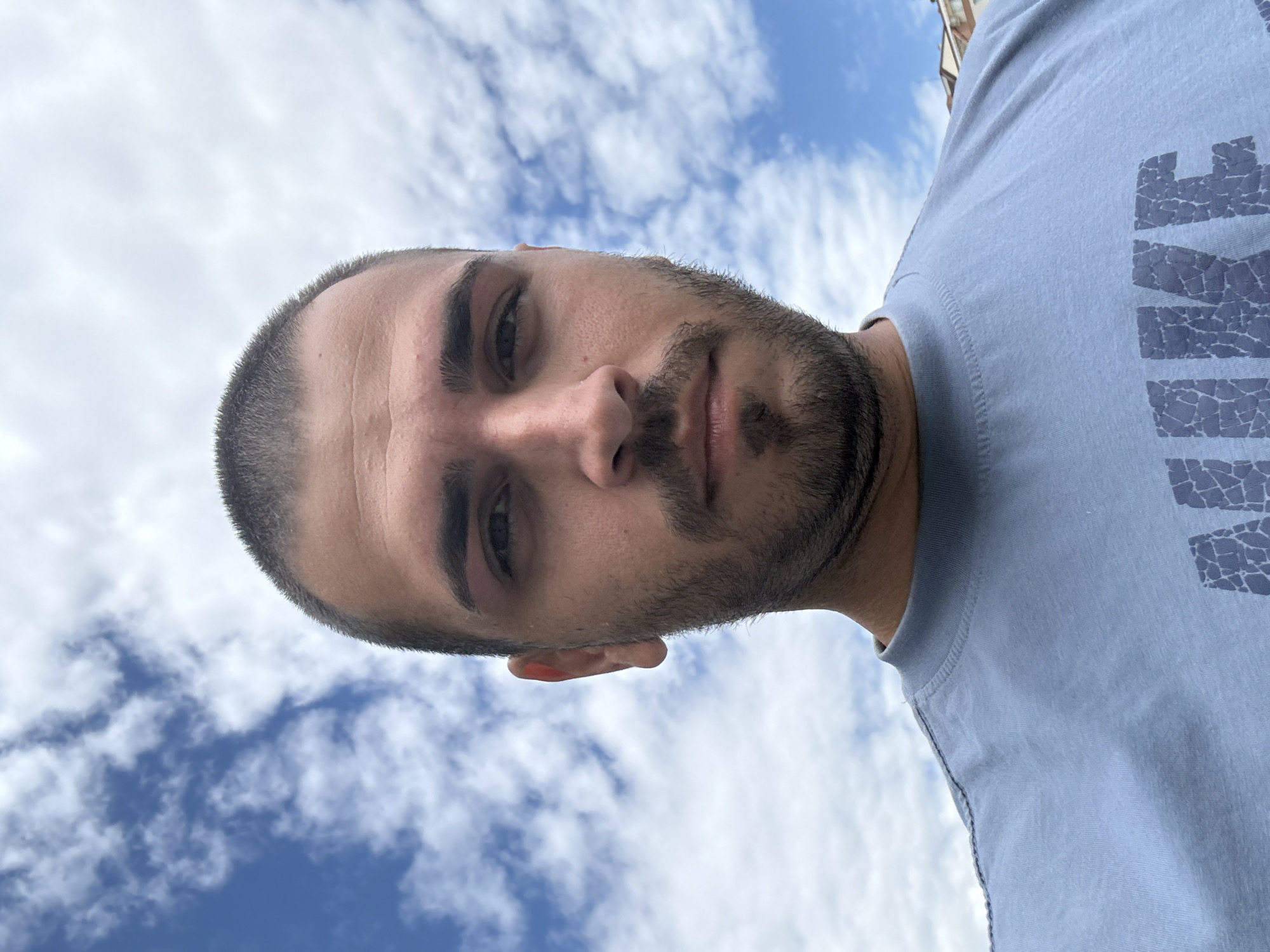 A low-angle close-up photograph shows a Caucasian male outdoors against a backdrop of blue sky with scattered clouds. He has a short, dark, closely-cropped hairstyle and a trimmed beard. He is wearing a light blue t-shirt with a large "NIKE" graphic on the right side of the frame. The location is indeterminate, but the sky suggests the photograph was taken outdoors. No evidence indicates the location is in Burgas, Bulgaria.