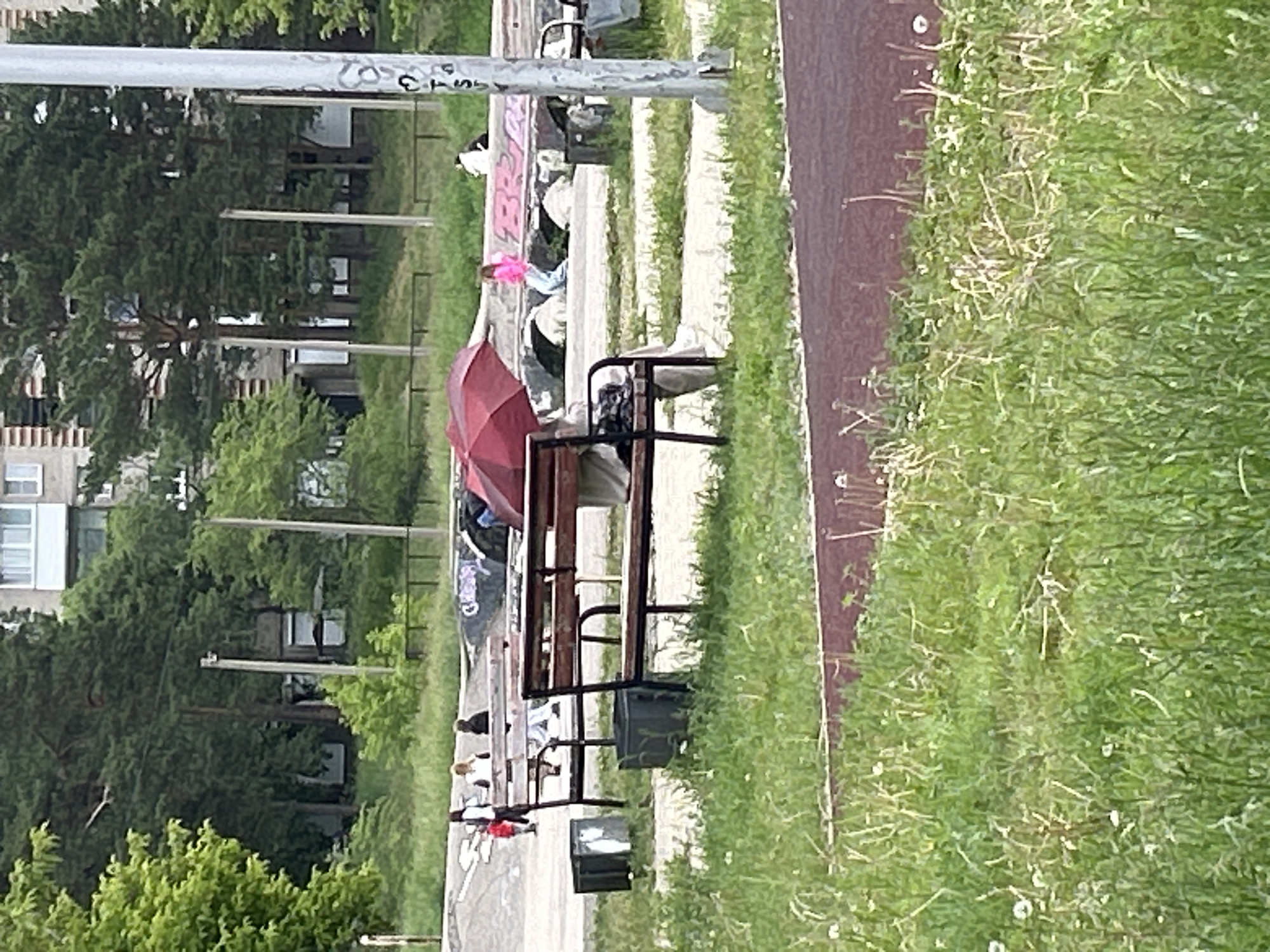 The image depicts a public park area in Angarsk, Russia, likely during daytime given the ambient lighting. In the foreground, a wooden bench is occupied by a person holding a red umbrella over their head. Two dark trash receptacles are positioned near the bench. The middle ground reveals a skate park with concrete structures and graffiti. Several individuals are visible in this area, some standing, others walking. One person is wearing a bright pink top and blue pants. The background features a multi-story building with a light-colored facade and visible windows. Dense green trees surround the park and building. The ground is covered with grass and a reddish-brown path. A metal pole with some markings is present on the right side of the image.