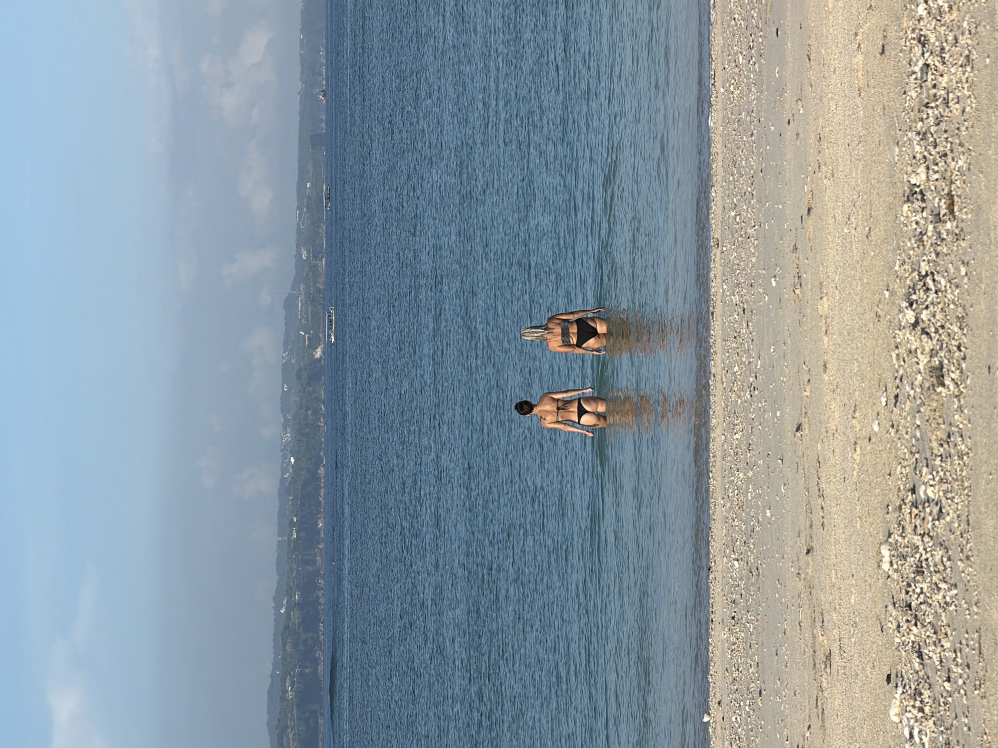 The image shows two individuals in a body of water, likely the ocean, adjacent to a beach. The location is not Kuta, Indonesia. One individual has blonde hair and is positioned slightly further from the shore than the other. Both individuals are wearing dark-colored swimwear. The beach is composed of sand and small stones or shells. The water is blue, and the sky is partially cloudy. A landmass with trees is visible in the distance.