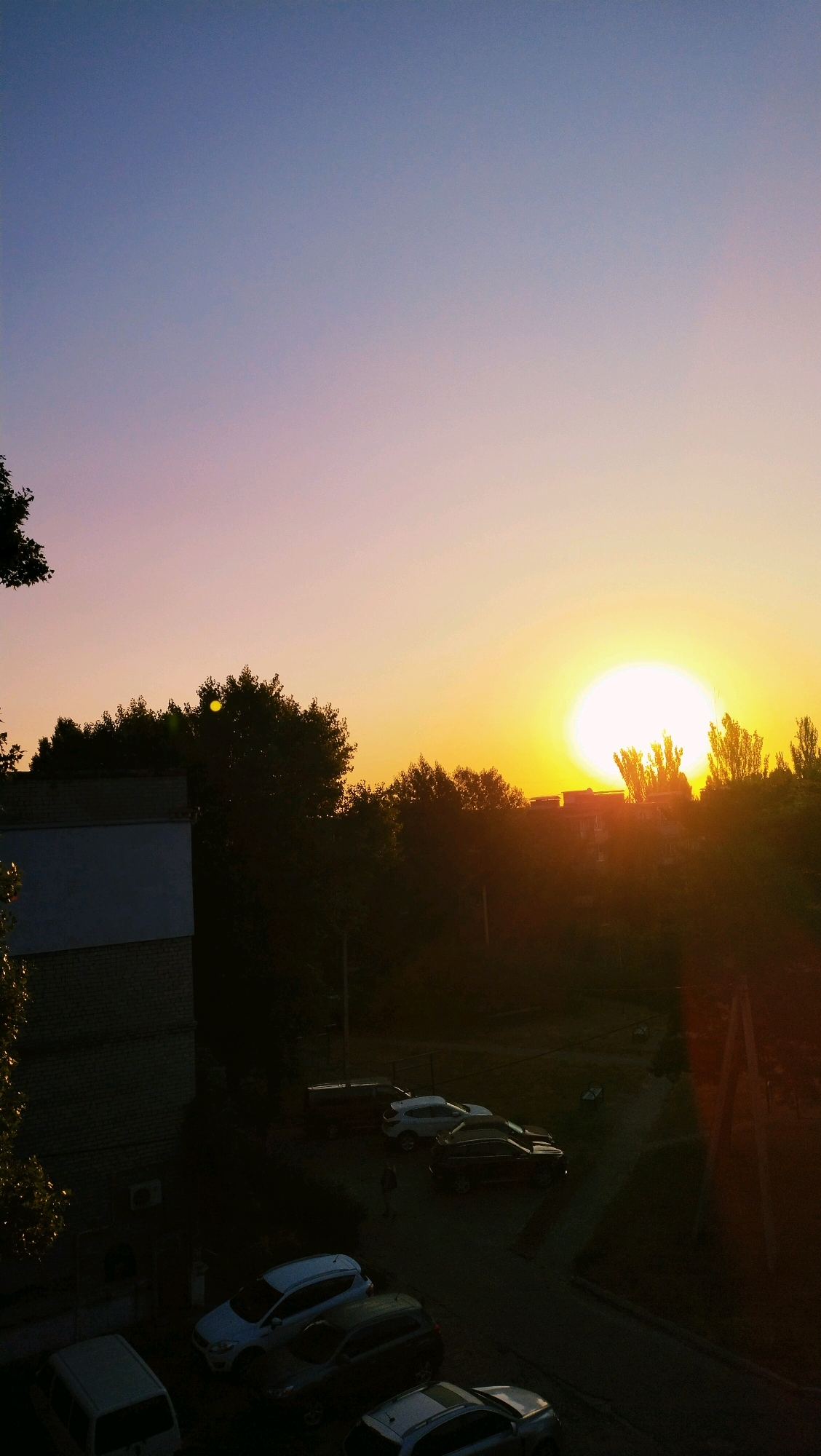 This image captures a sunset over a residential area in Horishni Plavni, Ukraine. The sky transitions from a deep blue at the top to soft shades of pink and orange as it nears the horizon. A large, bright sun is setting behind a line of trees and apartment buildings. In the foreground, a multi-story apartment building is visible on the left, casting a shadow over a parking lot. Several cars are parked in the lot, including a white SUV, a dark sedan, and a white hatchback. The ground appears to be asphalt. The overall scene is tranquil, with the warm glow of the sunset illuminating the urban landscape.