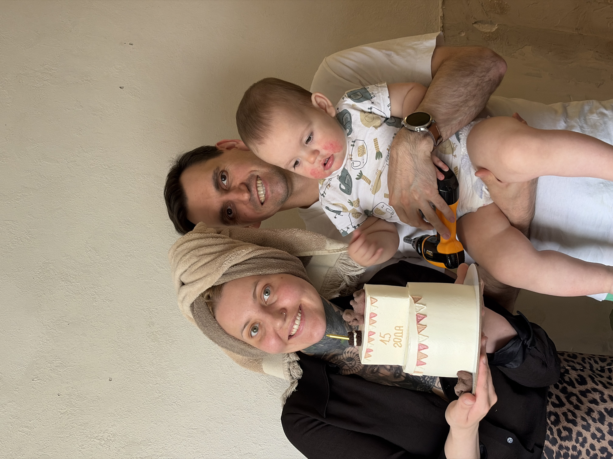 A family portrait consisting of a woman, a man, and a baby, possibly taken indoors in a residence within the city of Chaikovsky, Russia. The woman is in the foreground, wearing a dark top and a towel wrapped around her head. Visible tattoos are present on her neck and chest. She is holding a multi-tiered cake decorated with small pink triangles and gold lettering that reads "4.5 20.0A" (likely indicating a birthday celebration). A lit candle is placed on top of the cake. The man is positioned behind the woman, smiling and holding the baby. The baby is wearing a onesie with animal patterns and has some redness on its face. The man is also holding a yellow and black power drill near the baby. The backdrop consists of a plain, light-colored wall.
