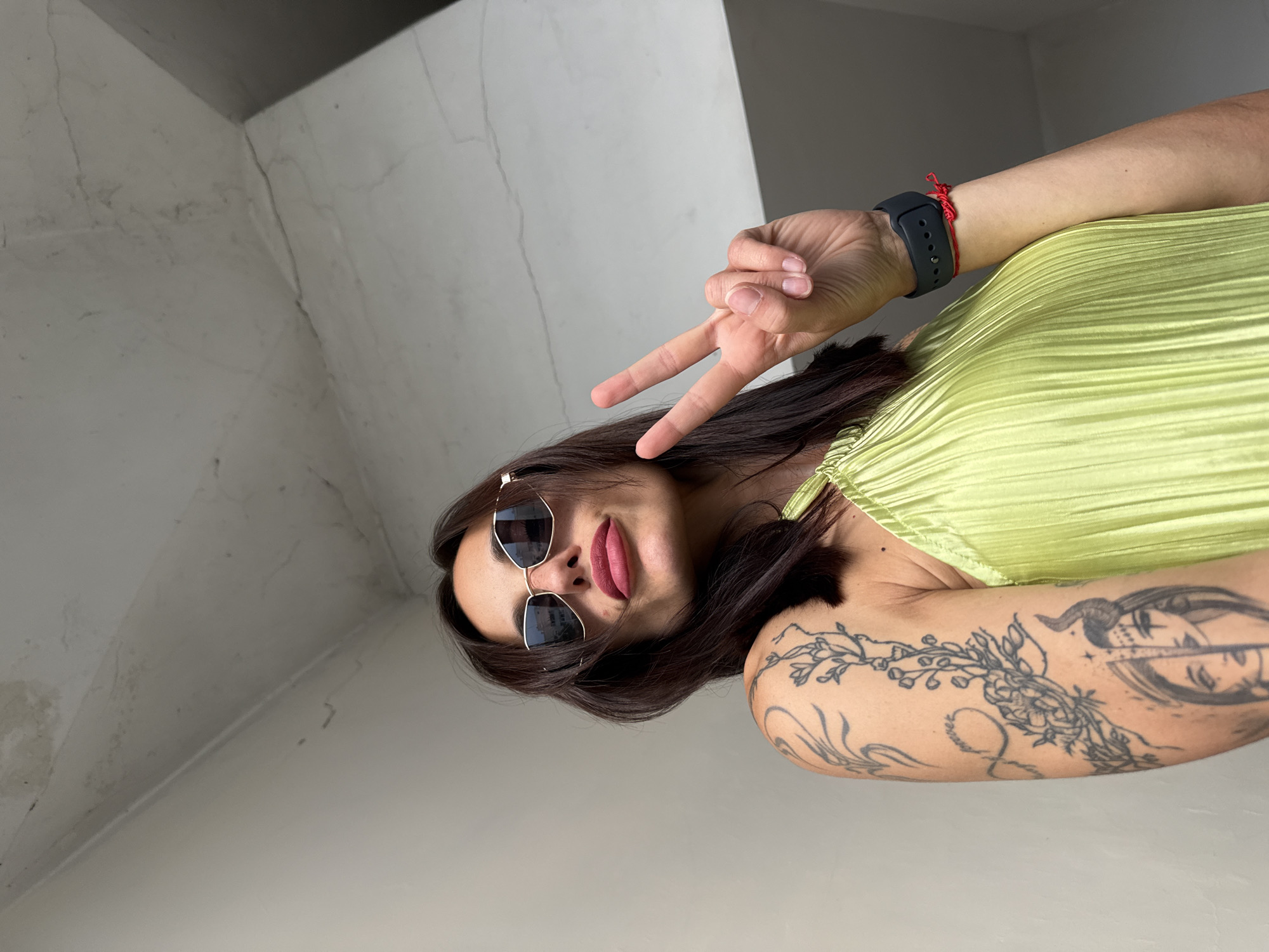 A woman wearing sunglasses and a lime green pleated dress poses for a selfie, holding up a peace sign with her right hand. She has a tattoo on her left arm. A dark smartwatch with a red string bracelet is visible on her right wrist. The background consists of a concrete wall and ceiling. The photo was taken in Pleven, Bulgaria.