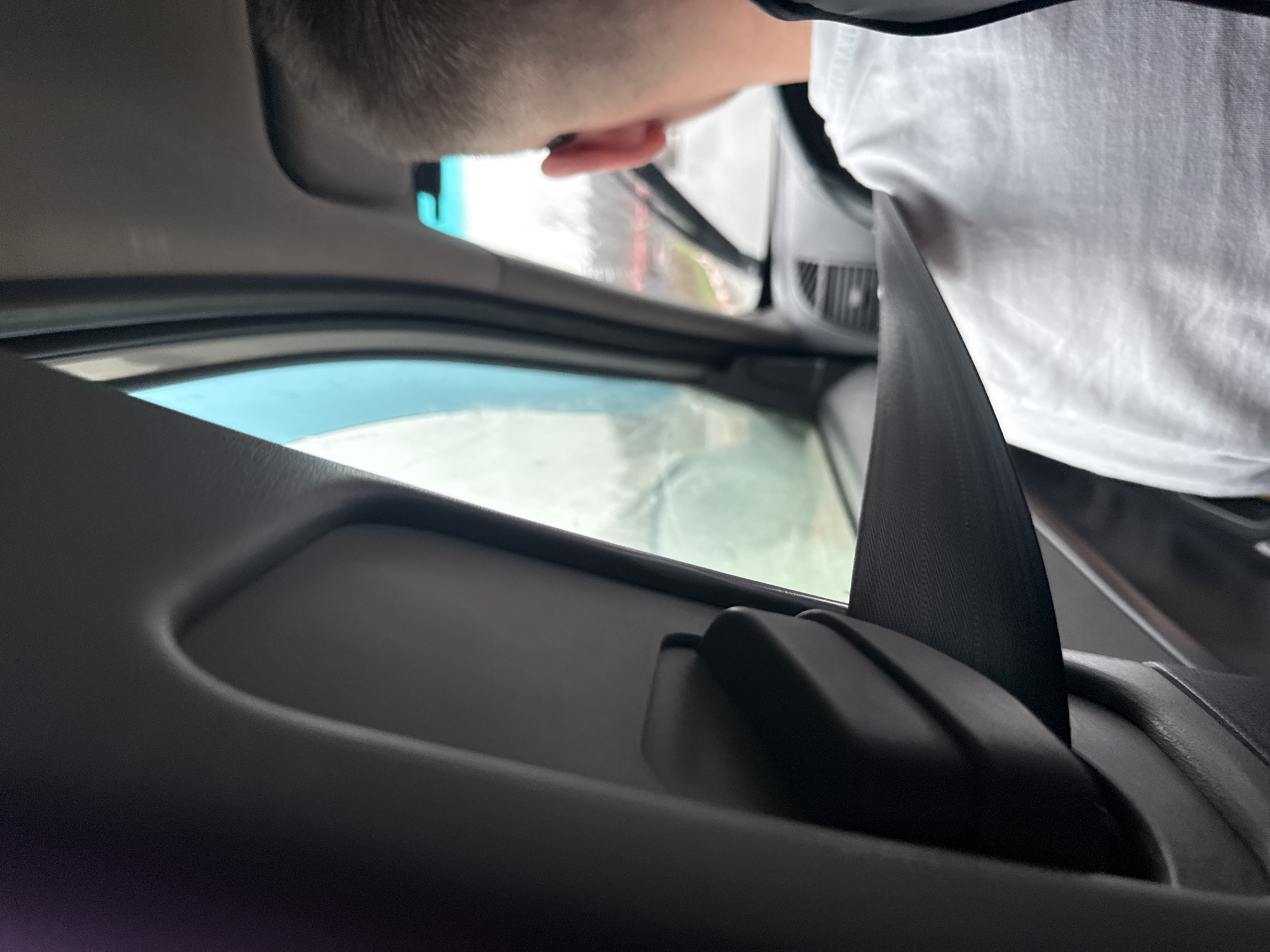 This image captures the interior of a vehicle, likely a car or van, viewed from a low angle, possibly from a rear seat looking towards the front passenger side. In the foreground, a prominent black seatbelt stretches diagonally across the lower right. A person, seen from the back of their head with short hair and a visible ear, occupies the upper part of the frame, wearing a white t-shirt.
The person appears to be positioned near the vehicle's front side, possibly leaning forward or looking out the side window. The car's grey interior door panel and window frame are visible, with the window showing a blurred view of the outside. The exterior scene is indistinct, suggesting either motion or overcast weather conditions during the daytime. A light blue tint, possibly from a mirror or part of the vehicle's structure, is visible near the person's head. No specific activity beyond observing the outside is clearly discernible. There are no visible texts in the image.