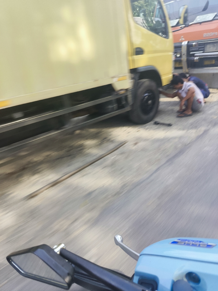 The image shows a yellow truck parked on the side of a paved road in Sukatani, Indonesia (latitude: -6.617, longitude: 107.406). Two men are crouched down next to the truck, working on a tire. The man closest to the camera is holding a wrench and appears to be tightening a lug nut on the tire. The truck is in the foreground of the image, and there is an orange truck parked behind it. In the foreground, the rear-view mirror of a light blue scooter can be seen, indicating the image was taken from a moving scooter. The road is in good condition and the day is clear. The scene is typical of roadside vehicle repair in an urban setting.