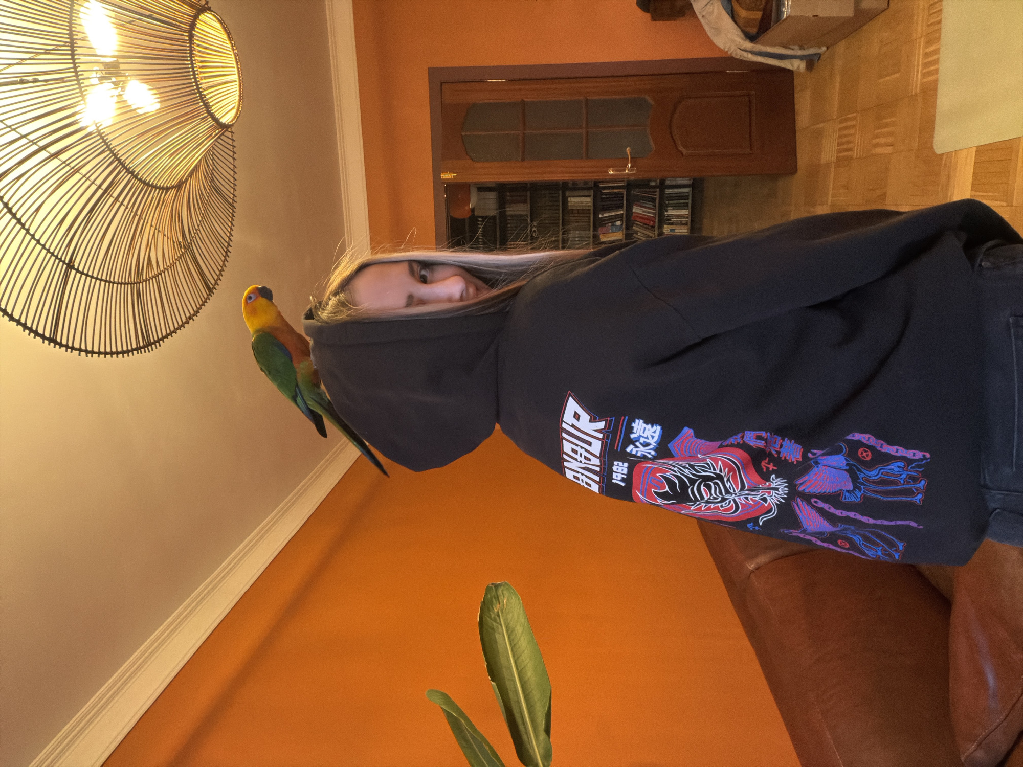 A person wearing a dark hooded sweatshirt with colorful Japanese-inspired graphics stands indoors. A Sun Conure parrot is perched on the person's left shoulder. The background includes an orange wall, a large wicker light fixture, a brown wooden door, and what appears to be a shelf with books or DVDs. A plant is visible in the foreground. The location, Khimki, Russia, cannot be determined from the image.