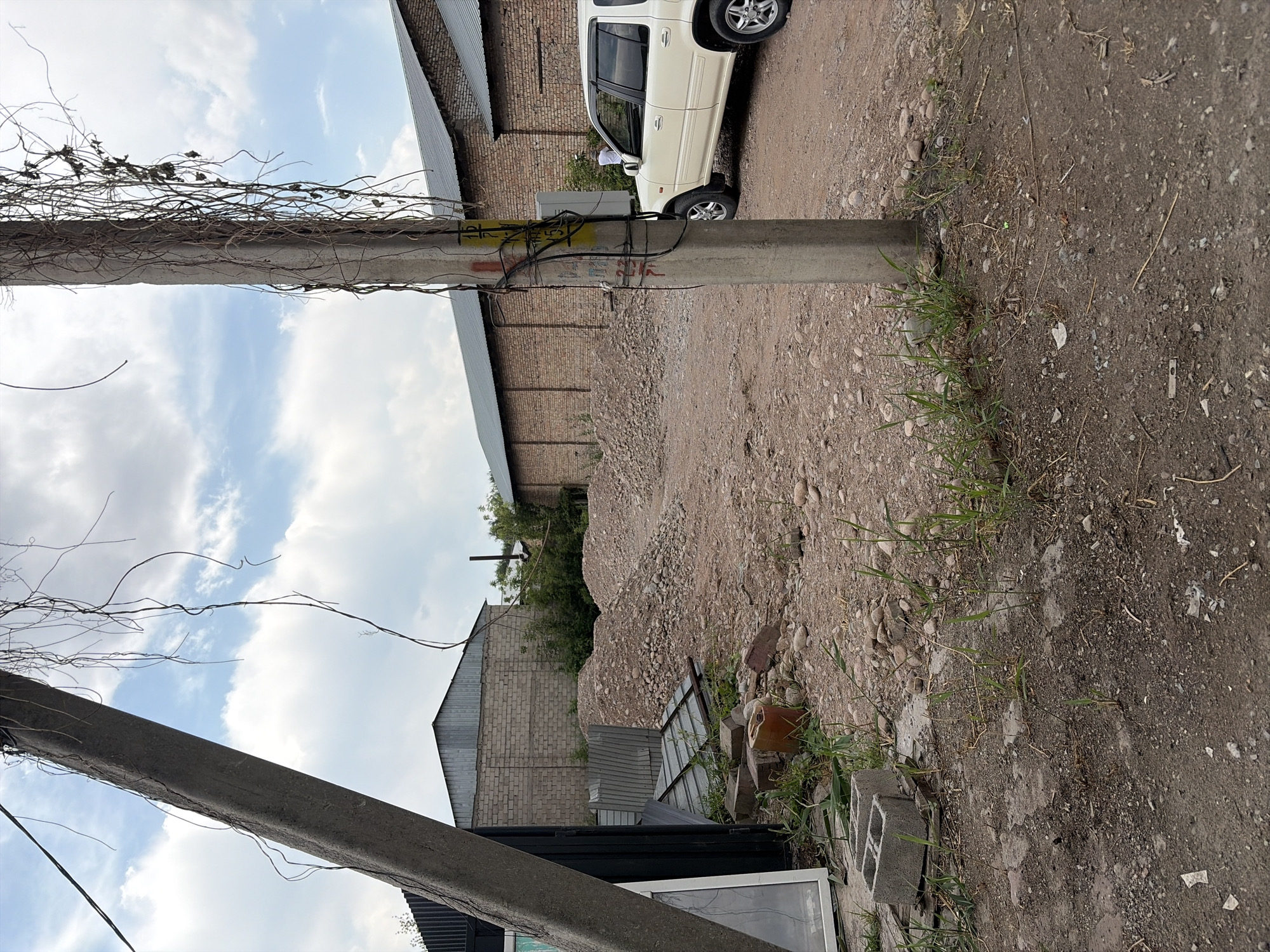 The image depicts an outdoor scene in the city of Kant, Kyrgyzstan, characterized by a construction or demolition site. In the foreground, a concrete utility pole stands prominently, adorned with numerous wires and some dried, trailing vines. To the left, a large, angled concrete pillar is visible, along with parts of a dark, rectangular structure that appears to be a building or shed with a large window. The ground is a mixture of dirt, gravel, and scattered debris, including concrete blocks, bricks, and metal sheeting.
In the mid-ground, large mounds of rubble and small stones dominate the landscape, suggesting excavation or demolition activities. Behind the rubble, several long, low-rise brick buildings with corrugated metal roofs stretch across the scene. A white SUV is parked on the dirt road to the right of the utility pole, facing left. A person in a white shirt can be seen near the rear of the SUV, interacting with an electrical box. The sky above is partially cloudy, with a mix of blue sky and white clouds. The overall impression is one of an industrial or construction area in a state of disarray.