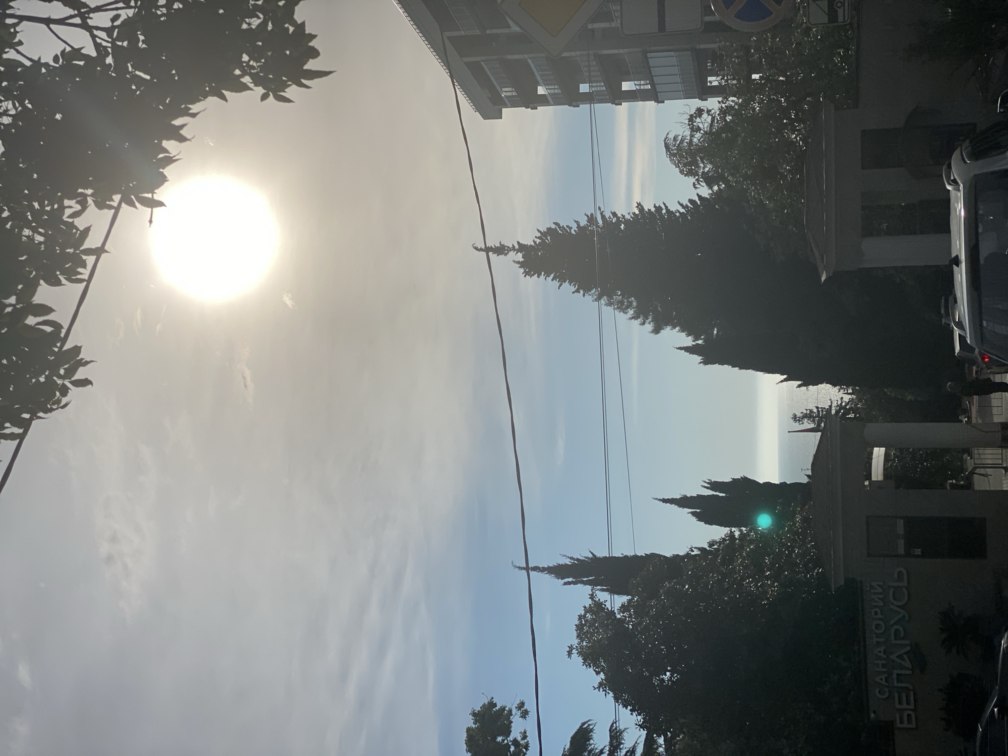 The image shows a street scene in Sochi, Russia, marked by the presence of the “Sanatorium Belarus” sign. The sky above is partly cloudy with a bright sun. Tall evergreen trees line the street, with overhead power lines visible. A building is partially visible on the left. To the right, a vehicle and pedestrian activity are apparent. The Black Sea is faintly visible in the distance.