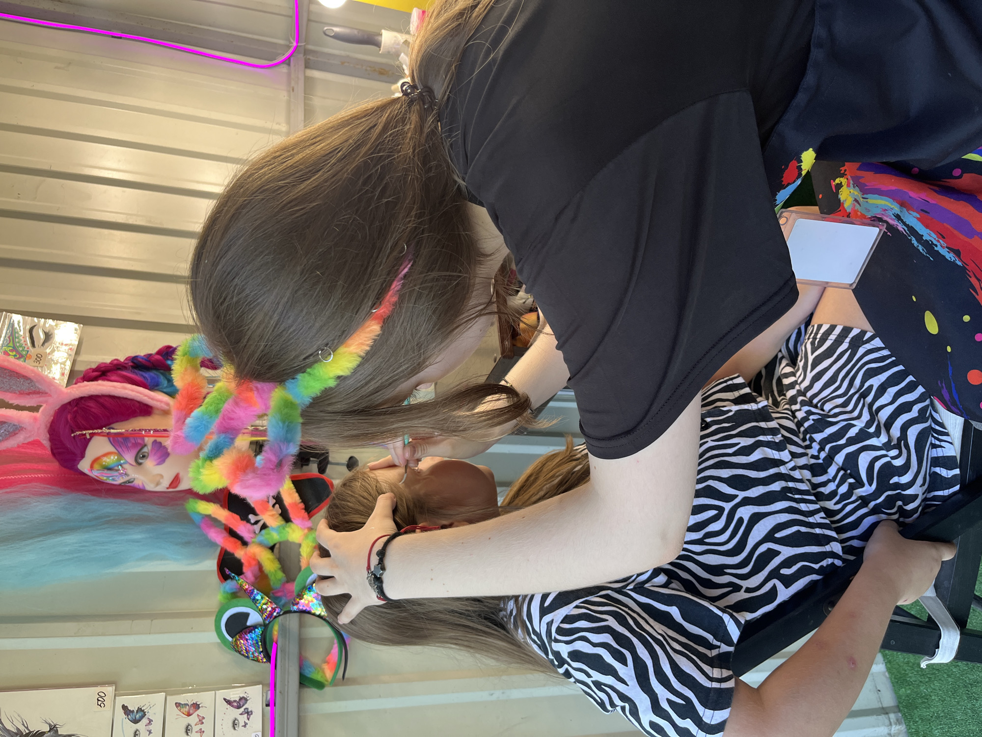 An outdoor stall is set up with a table covered in colorful accessories and face painting supplies. In the foreground, a young girl with long brown hair and wearing a black and white zebra-striped dress sits on a black chair. She is having her face painted by another person, whose hands and arm are visible in the frame. The painter is wearing a black t-shirt and a lanyard with an ID holder. The background shows a display of colorful wigs, bunny ears, and other party decorations, along with a corrugated metal wall.