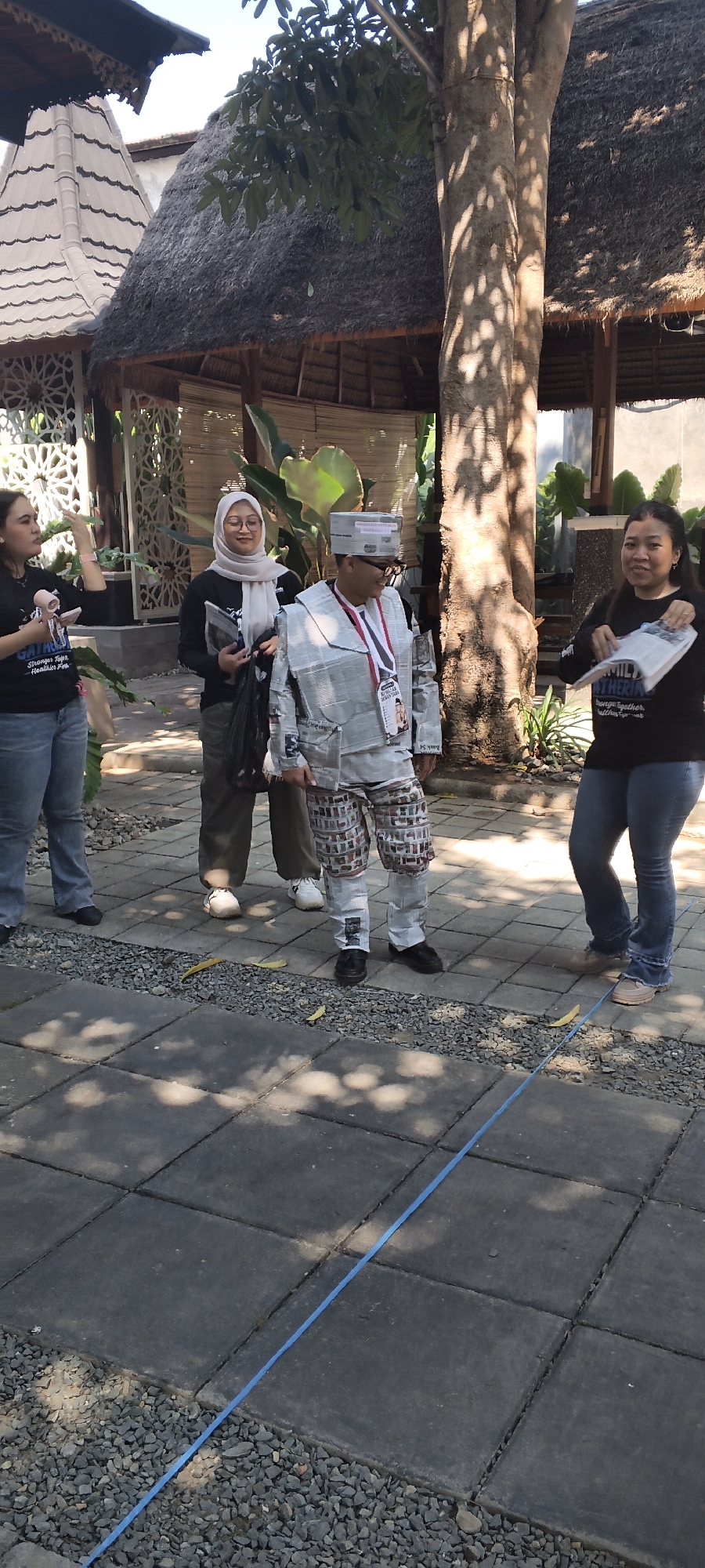 Four individuals are standing in an outdoor courtyard with paved stone flooring. To the left, a woman in a black t-shirt and blue jeans gestures with her right hand. Next to her, a woman wearing a light-colored hijab and a black t-shirt holds papers. In the center, a person dressed in a costume made of newspaper material, including a jacket, hat, and pants, stands with a medal around their neck. To the right, a woman in a black t-shirt and blue jeans holds papers and smiles. The background features a thatched-roof structure and a large tree. The scene is illuminated by natural light, casting shadows on the ground. This image was captured in Talise, Indonesia.