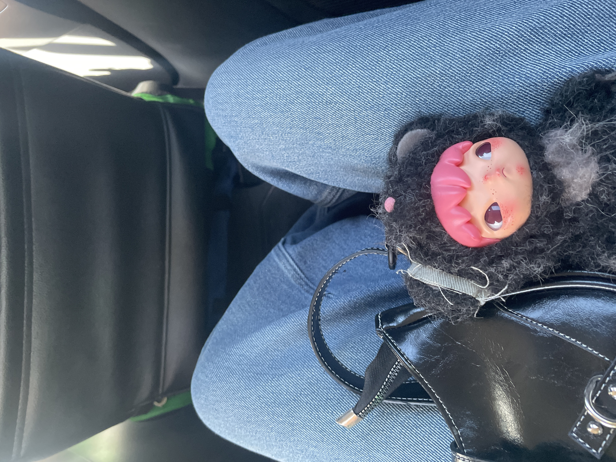 The image shows a person sitting in what appears to be a car. The person is wearing blue jeans. Resting on their lap is a black purse. On top of the purse is a small, black, furry doll with a pink face and pink hair. The inside of the car is visible, including part of the dashboard. The image does not provide enough information to confirm it was taken in Denpasar, Indonesia.