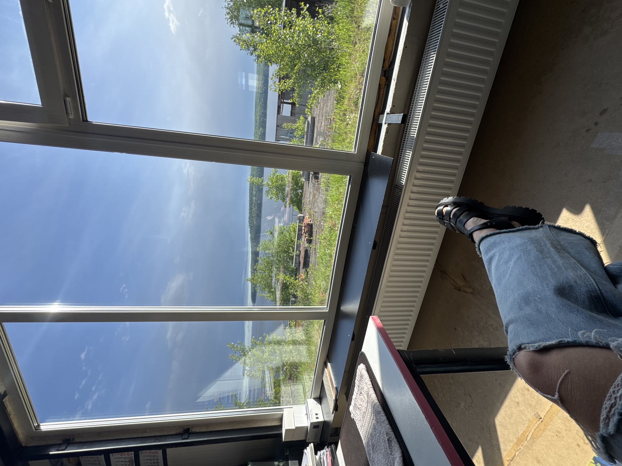 A person wearing blue ripped jeans and black sandals is seated inside a building, likely an apartment or house, looking out of a large multi-pane window. The view through the window features a landscape with a body of water, possibly a river or lake, bordered by trees and some structures. The sky is blue with scattered clouds, and the window reflects the sky, adding to the bright and open feel of the scene. Inside, a white radiator runs along the wall next to the window. The lower portion of the image shows part of a desk or table with items such as books or paperwork. The location is Chaikovsky, Russia.