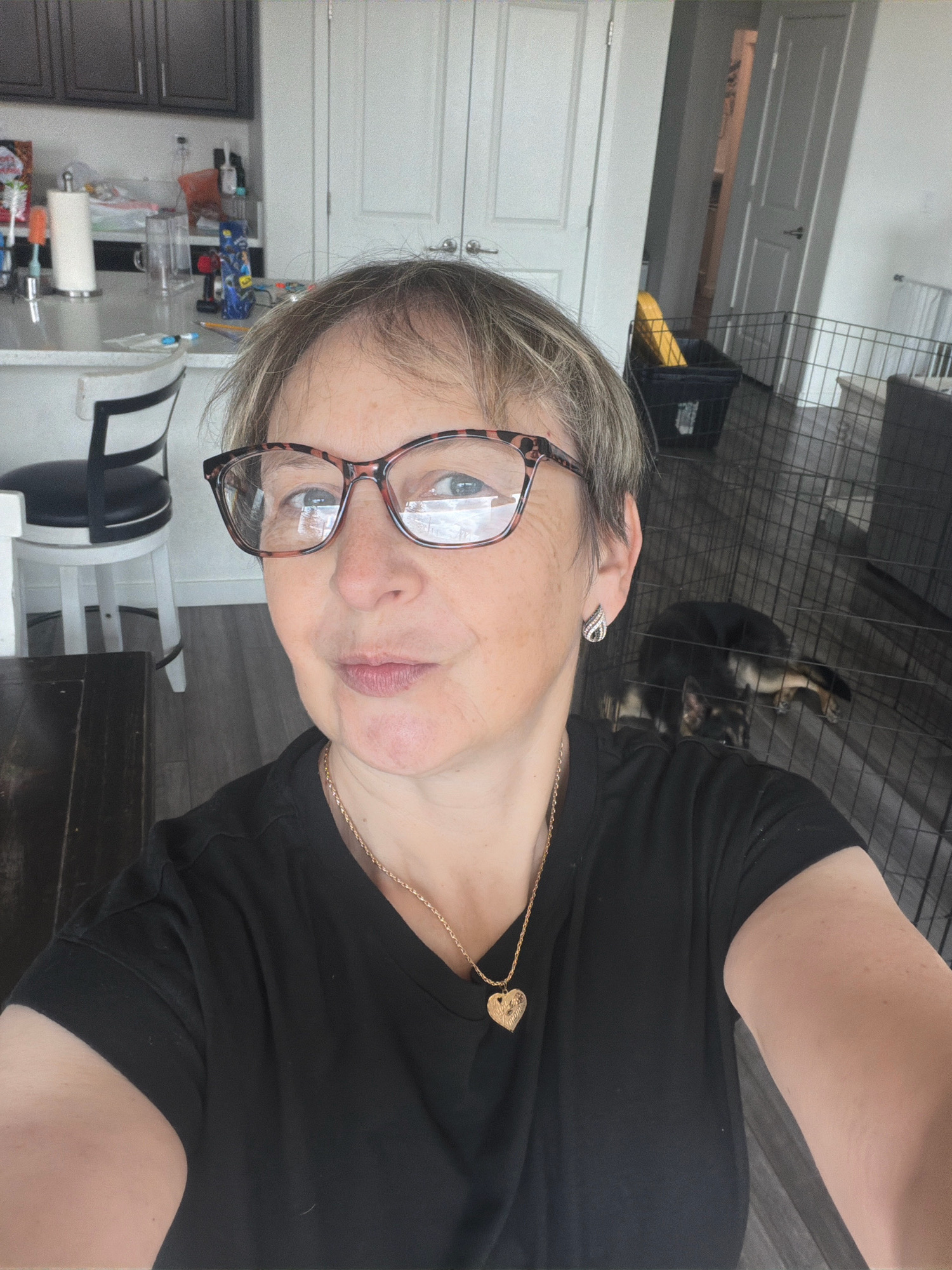 A medium shot captures a woman with short, blonde hair and glasses, wearing a black t-shirt and a gold necklace with a heart pendant, taking a selfie in an indoor setting that appears to be a home. She is looking directly at the camera with a neutral expression. In the background, a kitchen counter with various items is visible, as well as a dog in a cage. The flooring is a dark wood laminate. The overall lighting suggests it is daytime.