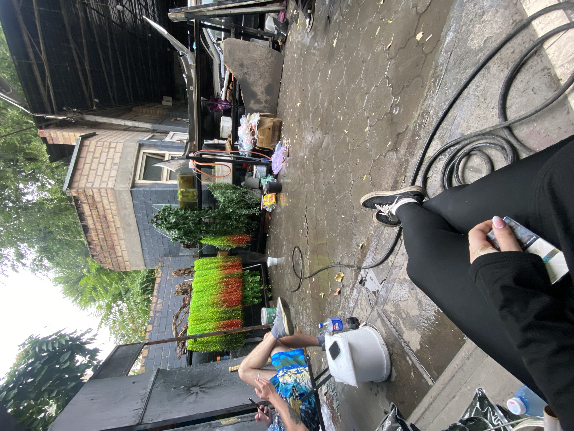 The image depicts an outdoor area, possibly a backyard or workshop, with a wet, paved ground suggestive of recent rain. In the foreground, a person's legs are visible, wearing black leggings and sneakers, holding a cigarette pack. Another person is seated in a folding chair on the left, holding a phone and a cigarette, dressed in shorts and sneakers.
Behind them, there are various objects and structures. A vibrant green and orange hanging plant display is prominent. There are also storage shelves with items like boxes, plastic bags, and what appears to be car parts. A partially visible building with brick and dark-colored walls forms the background. The scene is cluttered, with some debris scattered on the ground. The presence of a hose and what looks like a water dispenser suggests a functional or work-related space. The overall environment appears somewhat unkempt. The location is identified as Bishkek City, Kyrgyzstan.