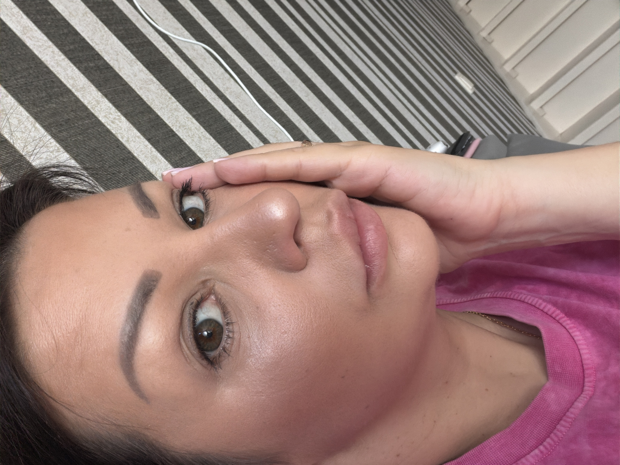 The image shows a close-up of a woman's face, positioned on the right side of the frame. She has light brown eyes, dark brown hair pulled back, and is wearing a bright pink t-shirt. Her right hand is placed on her cheek, with fingernails painted light pink. The background consists of a striped pattern of dark grey and beige, with a white cord visible on the right. The image appears to be taken indoors. There is no indication of the location being the city of Sochi, Russia.
