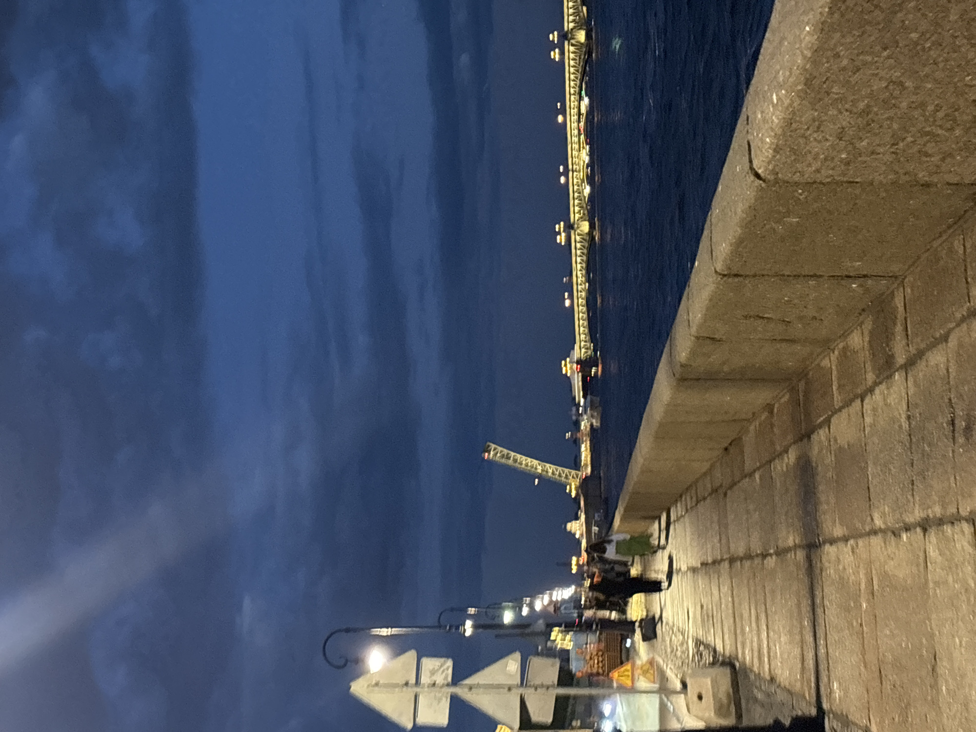 The image captures a twilight scene along the Neva River embankment in Saint Petersburg, Russia. A stone walkway, paved with large rectangular stones, runs along the foreground, bordered by a thick stone embankment wall on the right. Several lampposts, featuring ornate curved arms, are spaced along the walkway, providing illumination. Two figures are walking away from the camera on the left side of the walkway.
In the background, a wide river, likely the Neva, is visible, its surface rippled by a gentle breeze. Across the river, a long bridge is illuminated with a series of lights. To the left of the bridge, a drawbridge is raised, its structure forming a steep angle against the darkening sky. The sky itself is a deep blue, with clouds casting shadows and hinting at the lingering light of dusk.
The overall impression is one of a serene evening on a prominent Saint Petersburg landmark, with the city's iconic bridges and waterfront setting clearly depicted.
