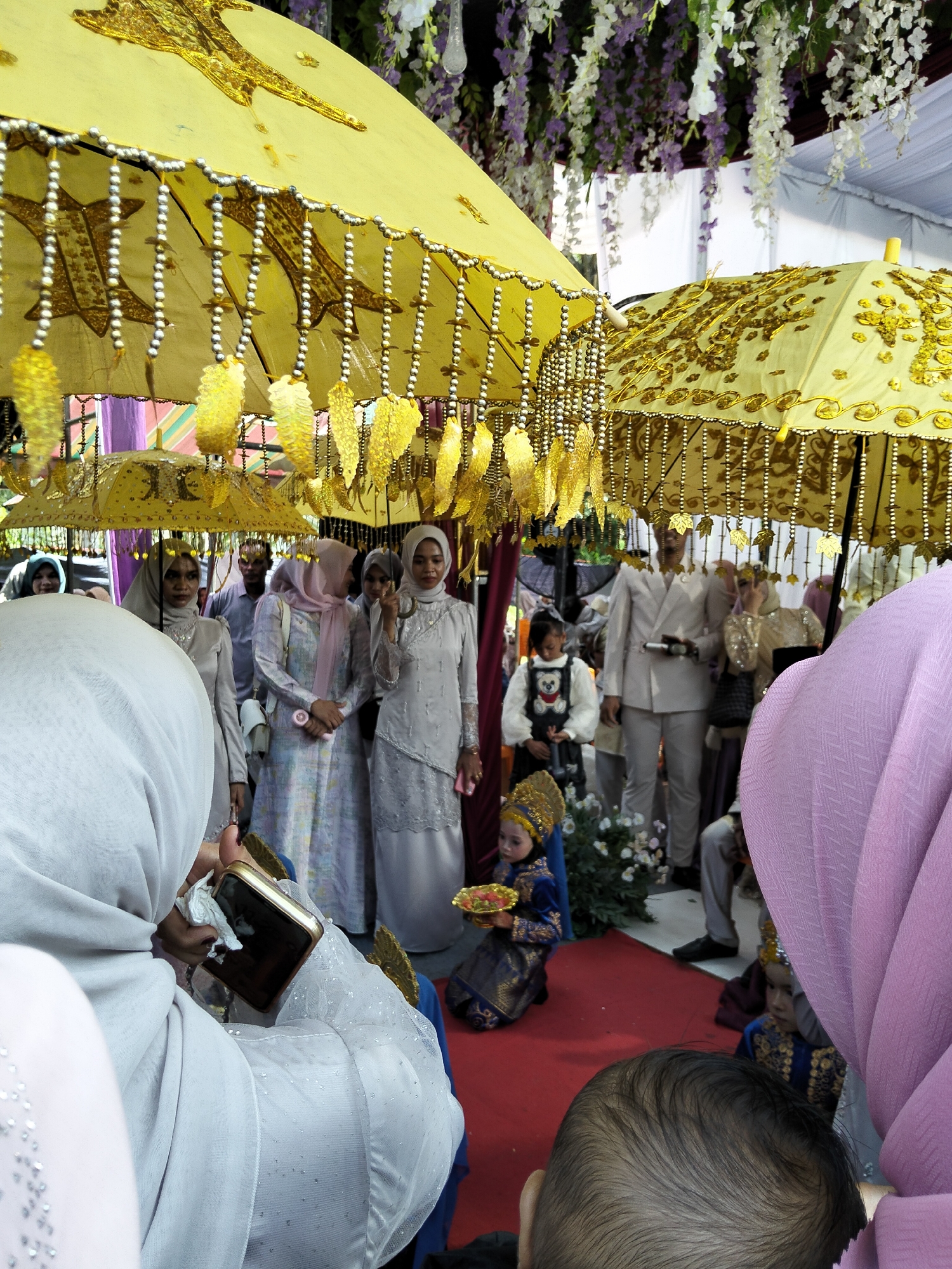 In an outdoor event tent adorned with hanging flowers, likely in Pulonga, Indonesia, several people are gathered under large, ornate yellow umbrellas decorated with gold sequins and beads. In the foreground, a person wearing a white hijab is holding a smartphone. To their left, a woman in a silver hijab and dress is visible. Further back, a group of women in various hijabs and traditional attire are present, some holding umbrellas. A man in a light-colored suit stands behind them. In the middle ground, two young children in traditional clothing are seated on a red carpet, one holding a small plate of fruit. The overall scene suggests a cultural ceremony or celebration.