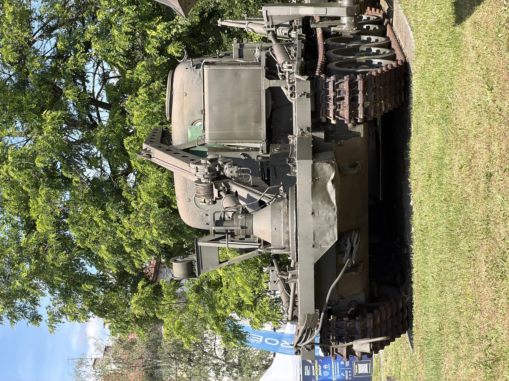 In Sofia, Bulgaria, on a sunny day, a large, olive-drab, tracked military vehicle is displayed outdoors on a grassy lawn. The vehicle, likely a recovery or engineering vehicle, features a robust construction with visible tracks, multiple wheels, a large winch mechanism at the front, and a boom or crane arm. There is a blue banner with white text on the left side of the image, partially obscuring the front of the vehicle. The background is dominated by lush green trees and a clear blue sky with some scattered clouds. The overall scene suggests an outdoor exhibition or event.