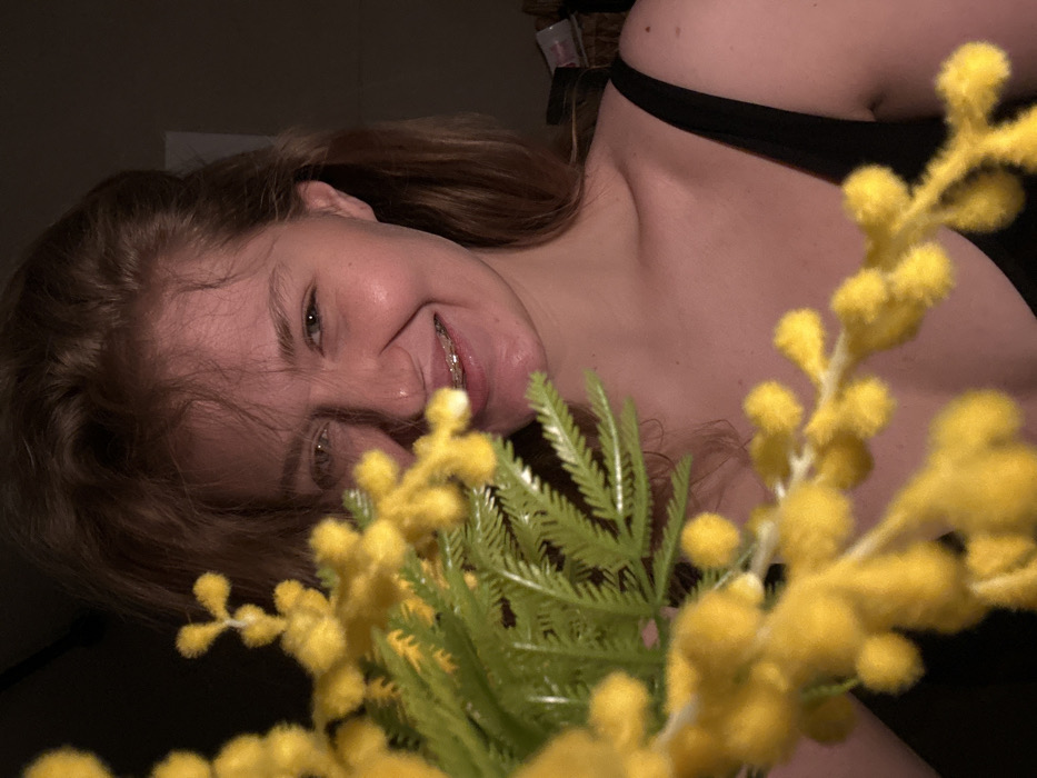 A close-up photograph depicts a smiling female subject partially obscured by a foreground bouquet of out-of-focus yellow mimosa-like flowers with green foliage. The subject has light hair and is looking directly at the camera, revealing visible dental braces on her upper teeth. She wears a dark-colored strap garment, likely a tank top or camisole, visible on her right shoulder and upper chest. The background is dark and indistinct, indicating an indoor, dimly lit environment. There are no identifiable visual cues within the image to specify a location in the city of Kazan, Russia.
