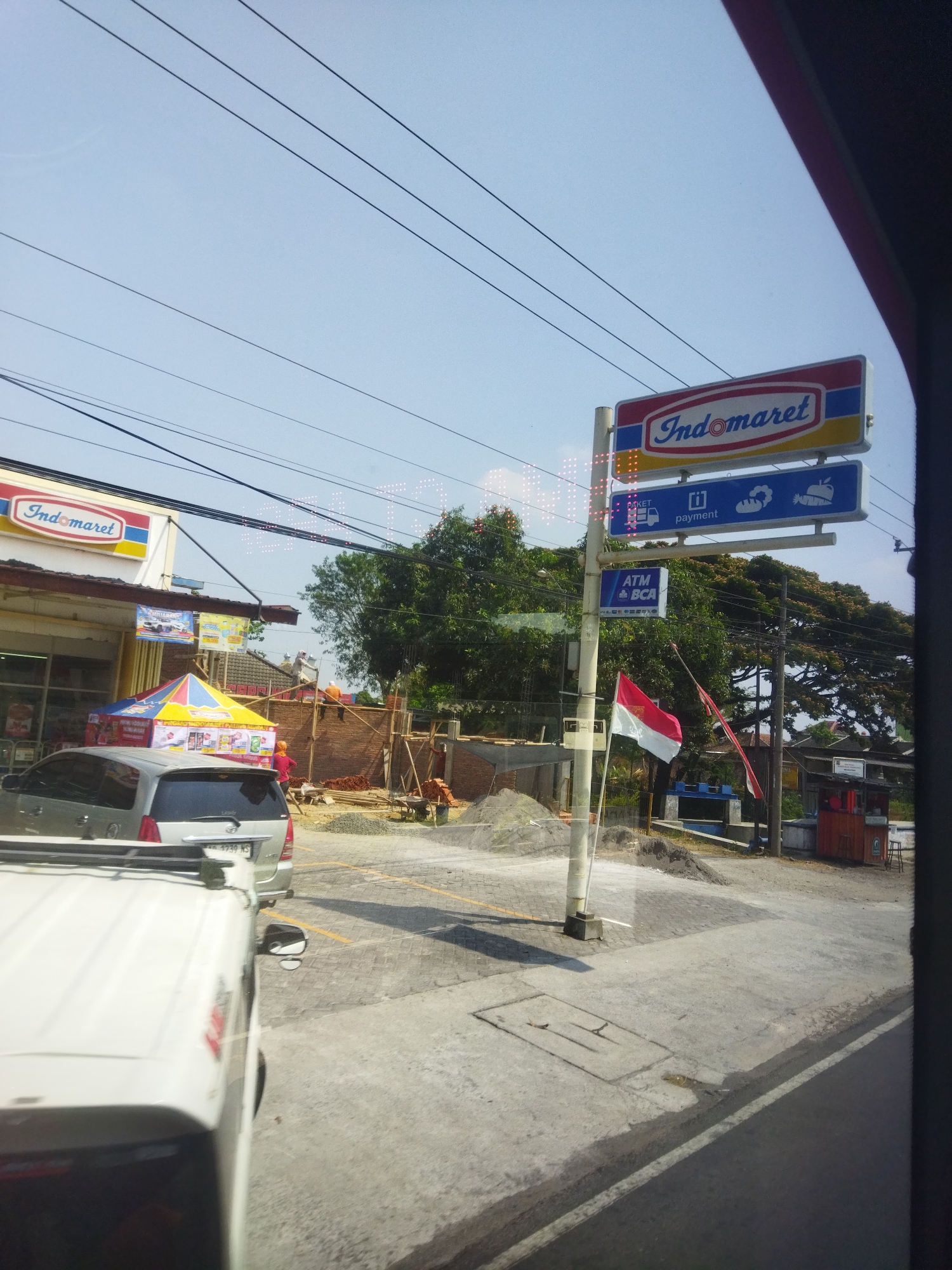 This image captures a street scene in Bulakrejo, Indonesia, on a sunny day. On the left, a row of buildings includes an Indomaret convenience store with its distinctive signage. In front of the store, a tent is set up, possibly for promotions or as a vendor stall. A silver SUV is parked in front of the Indomaret.
To the right of the Indomaret, construction is underway, with workers in orange vests visible and a pile of bricks. A central pole holds multiple signs, including an Indomaret logo, a sign with "paket" and "payment" icons, and a sign for "ATM BCA." Two Indonesian flags, one larger than the other, are displayed, with the larger one hanging from a pole. Further to the right, a small red kiosk-like structure is visible, with a stool outside. The ground is paved, and a road with white markings runs through the scene. Power lines are strung across the clear blue sky. The image appears to be taken from inside a vehicle, indicated by the reflection on the glass and the partial view of the vehicle's interior in the foreground.