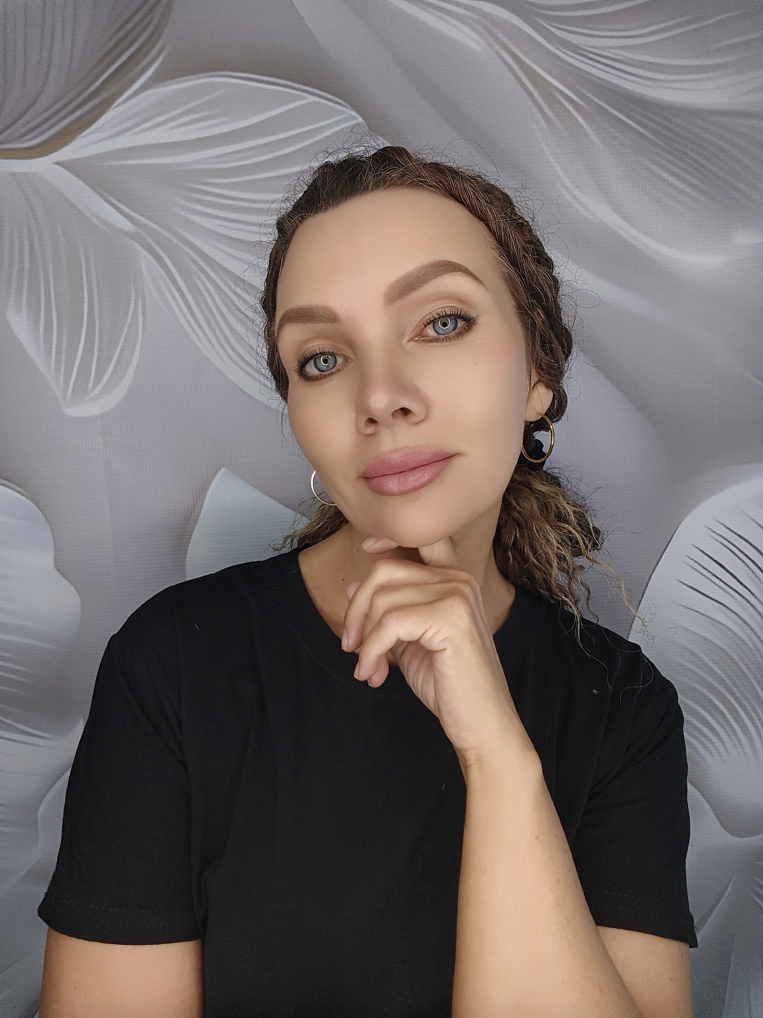 A medium shot shows a woman with blue eyes, light brown braided hair, and fair skin, wearing a black t-shirt. She is looking directly at the camera with her right hand on her chin, her left hand partially visible at her chest. The background is a grey textured wallpaper with white floral patterns. The image is taken in an indoor setting.