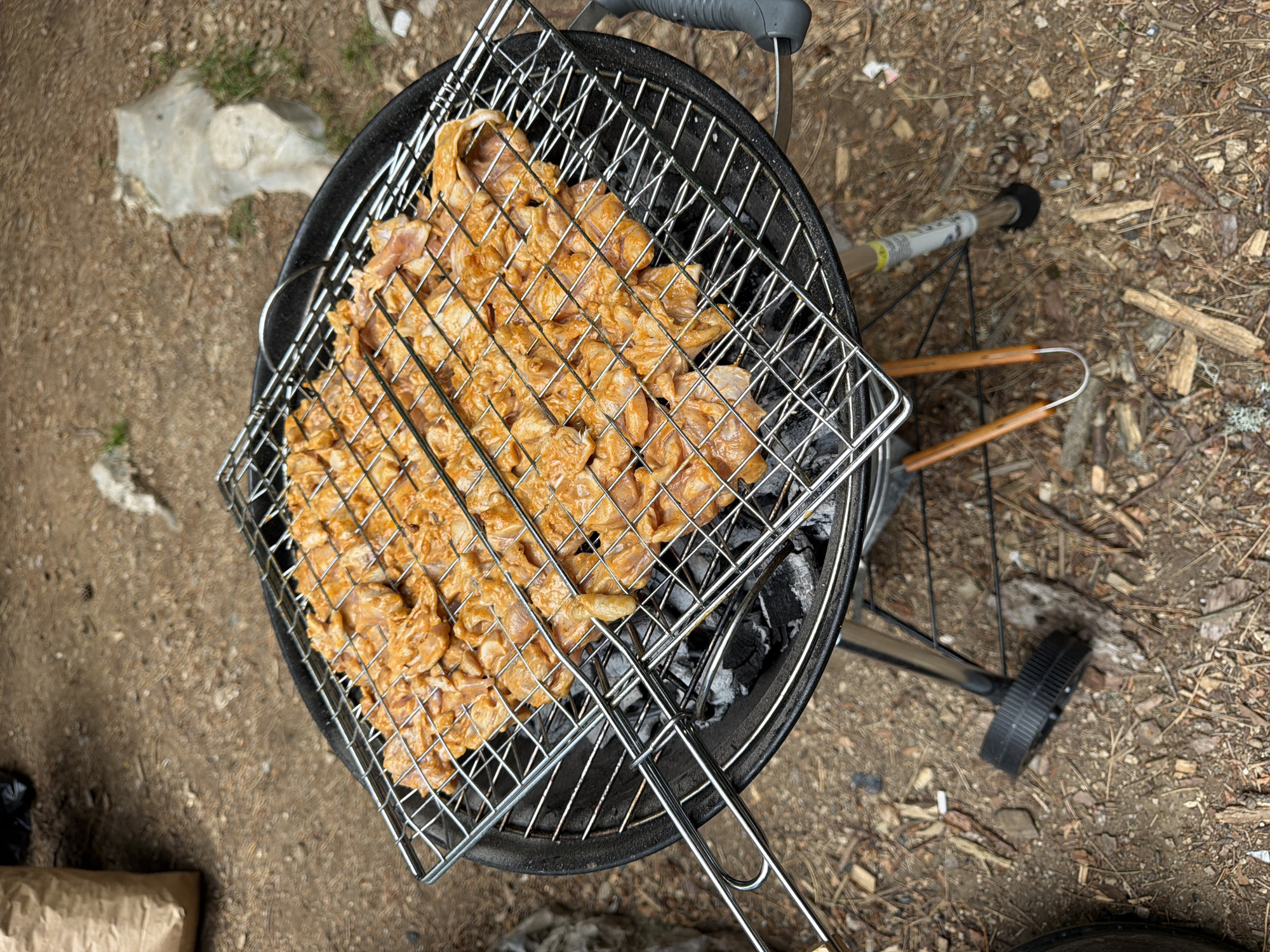 This overhead image captures an outdoor grilling scene in Mali Bostur, Montenegro, during the daytime. No individuals are visible.
The central subject is a black, round kettle-style charcoal grill. Placed on the grill is a rectangular metal cooking basket, filled with numerous pieces of raw, marinated meat, likely chicken or pork. The meat is covered in an orange-brown seasoning or sauce, indicating preparation for cooking. Below the cooking basket, glowing charcoal embers are visible, providing heat for the grilling process.
The setting is a natural, rustic outdoor area. The ground consists of dirt mixed with various natural elements such as pine needles, small pieces of wood, and sparse patches of grass. In the upper left portion of the image, a crumpled white or translucent plastic sheet or bag lies on the ground. Towards the bottom left, a portion of a brown paper bag is visible. The grill is equipped with wheels, one of which is partially visible on the right side. Nearby, a grilling tool, possibly tongs or a grate lifter with wooden handles, rests on the ground. A stick or pole with a white label and yellow tape is also on the ground to the right of the grill, but any text on it is unreadable. The scene conveys a relaxed, outdoor cooking activity in a natural environment.