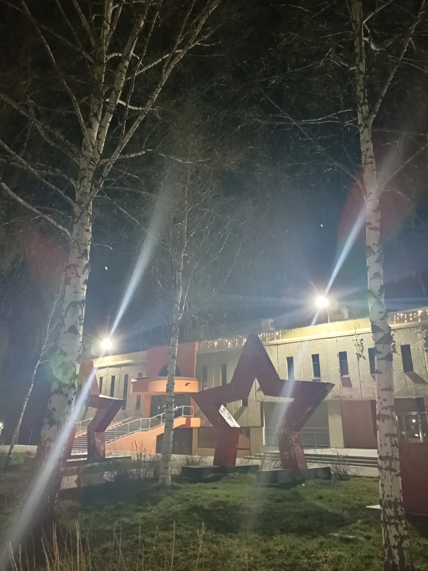 The image presents an outdoor night scene in Markovskii, Russia, featuring a building and prominent sculptures, framed by trees.
In the foreground and mid-ground, several birch trees with characteristic white, peeling bark and bare branches stand tall, reaching into the dark night sky. These leafless trees suggest a cold season, likely late autumn, winter, or early spring. Patches of green and dry grass cover the ground beneath them.
The central subject is a multi-story building, possibly a public or cultural institution, with a light-toned, possibly beige or yellowish, facade. It features numerous rectangular windows and distinctive orange-colored architectural elements, particularly around an external staircase leading to an entrance. Decorative string lights are visible along the building's roofline, providing additional illumination.
Positioned in front of the building are two large, red, five-pointed star sculptures. These stars appear solid and substantial, acting as significant landmarks or commemorative pieces.
The entire scene is brightly lit by artificial light sources, which produce noticeable lens flares and light beams across the image, particularly from the upper left and right, illuminating the trees and parts of the building. The sky is dark, indicating nighttime.
There are no people visible, and no specific activity or event is taking place, giving the scene a still and quiet atmosphere. The combination of the red stars and birch trees provides strong cultural and geographic cues consistent with the given Russian location. No visible text is present in the image.
