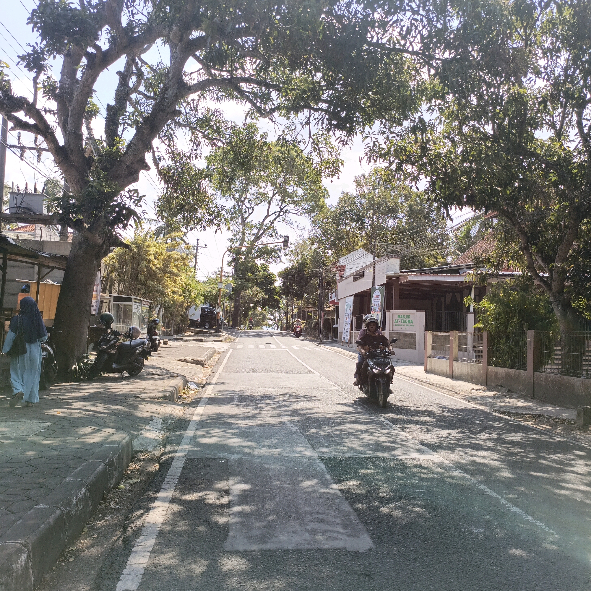 This image depicts a street scene in Salatiga, Indonesia, likely in a moderately developed area. The foreground features a paved road with a white dashed line marking the lane division. The road is partially shaded by dappled sunlight filtering through large trees lining the left side of the street. A pedestrian walkway, paved with interlocking blocks, runs along the left side of the road. On this walkway, a person wearing a light blue hijab and a matching long-sleeved top is walking away from the camera, carrying a black bag. To their left, parked motorcycles are visible, including two in the immediate vicinity of the pedestrian.
Further down the street, a motorcycle with two occupants, both wearing helmets, is traveling towards the camera. The driver is wearing a dark t-shirt, and the passenger is wearing a lighter-colored shirt. On the right side of the street, buildings are visible, including a structure with a white facade and a sign that reads "MASJID AT-TAQWA". A fence separates this building from the road. Trees with dense foliage are prevalent throughout the scene, creating a lush, green environment. Power lines are visible overhead, and traffic signals are present further down the road. In the background, more trees and buildings can be seen, suggesting a suburban or semi-urban setting. The overall atmosphere is that of a bright, sunny day.