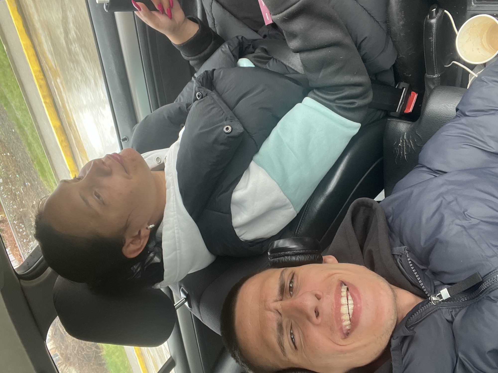 The image depicts two individuals inside a car. The person on the left is a woman with dark hair and earrings, wearing a white hoodie underneath a black and white jacket. The person on the right is a man with short hair, wearing a dark gray hoodie and a blue puffy jacket. Both are smiling. The interior of the car is visible, including the seats, seatbelts, and a cup of coffee. The outside environment appears to show a street with greenery and buildings. The city location is not identifiable from the image.