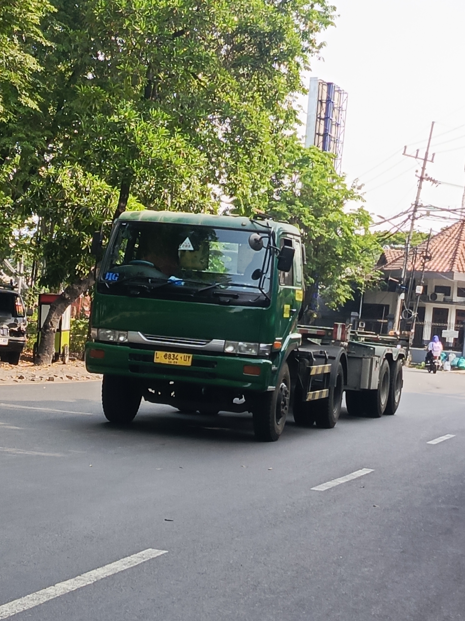 A green truck with the license plate L 8834 UY is seen driving on a paved road in Surabaya, Indonesia. The truck has a visible "BIG" sticker on its windshield and appears to be a flatbed truck. The surrounding environment includes lush green trees, buildings with tiled roofs, utility poles with wires, and a billboard structure in the background. The sky is bright and overcast. A motorcycle with a person wearing a purple headscarf is visible in the distance on the right side of the image, moving away from the truck. The road has white lane markings.