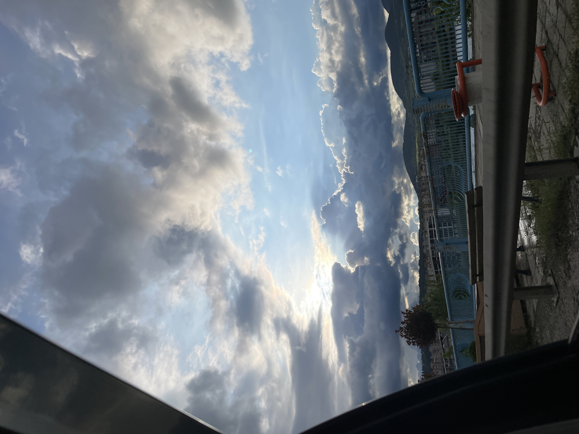 The image is a view of the sky with clouds, taken from within a vehicle in Kardzhali, Bulgaria. Visible through the sky are the sun and cumulus clouds of varying shades of gray and white. A portion of the bridge is visible on the right. The fence of the bridge is bluish. A railing with a lifebuoy sits on top of the bridge. There are trees and buildings near the bridge, but the scene is somewhat obscured by the clouds. The image appears to have been taken through a sunroof or window.