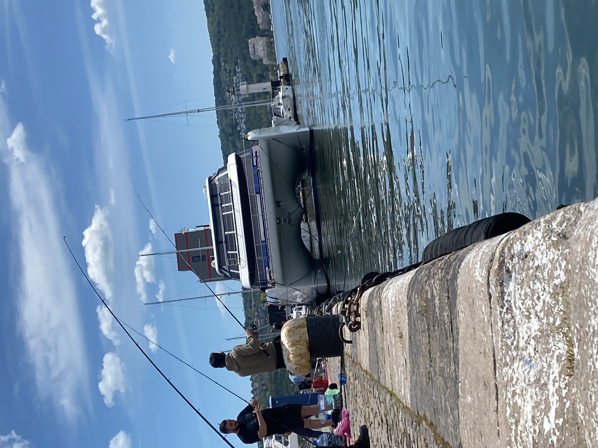Two men are fishing from a stone pier. A large boat is docked next to the pier. On the pier, there are cylindrical fenders, chains, and personal items. The water is still, reflecting the sky. In the background, there are buildings on a hillside. The location is Varna, Bulgaria.