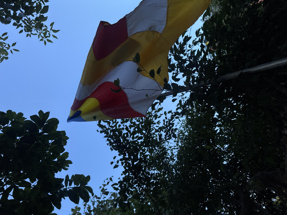 A Buddhist flag is depicted prominently, unfurled against a clear blue sky. The flag features five vertical stripes: blue, yellow, red, white, and orange, followed by a sixth vertical stripe composed of stacked horizontal sections of these five colors. It is attached to a dark grey flagpole, partially visible in the lower right and extending upwards. Dense, dark green foliage from multiple trees frames the upper left, lower left, and bottom right portions of the image, appearing as dark silhouettes due to backlighting. No individuals are present. The scene is situated within the outdoor environment of a Buddhist temple or pagoda in Nha Trang, Vietnam.