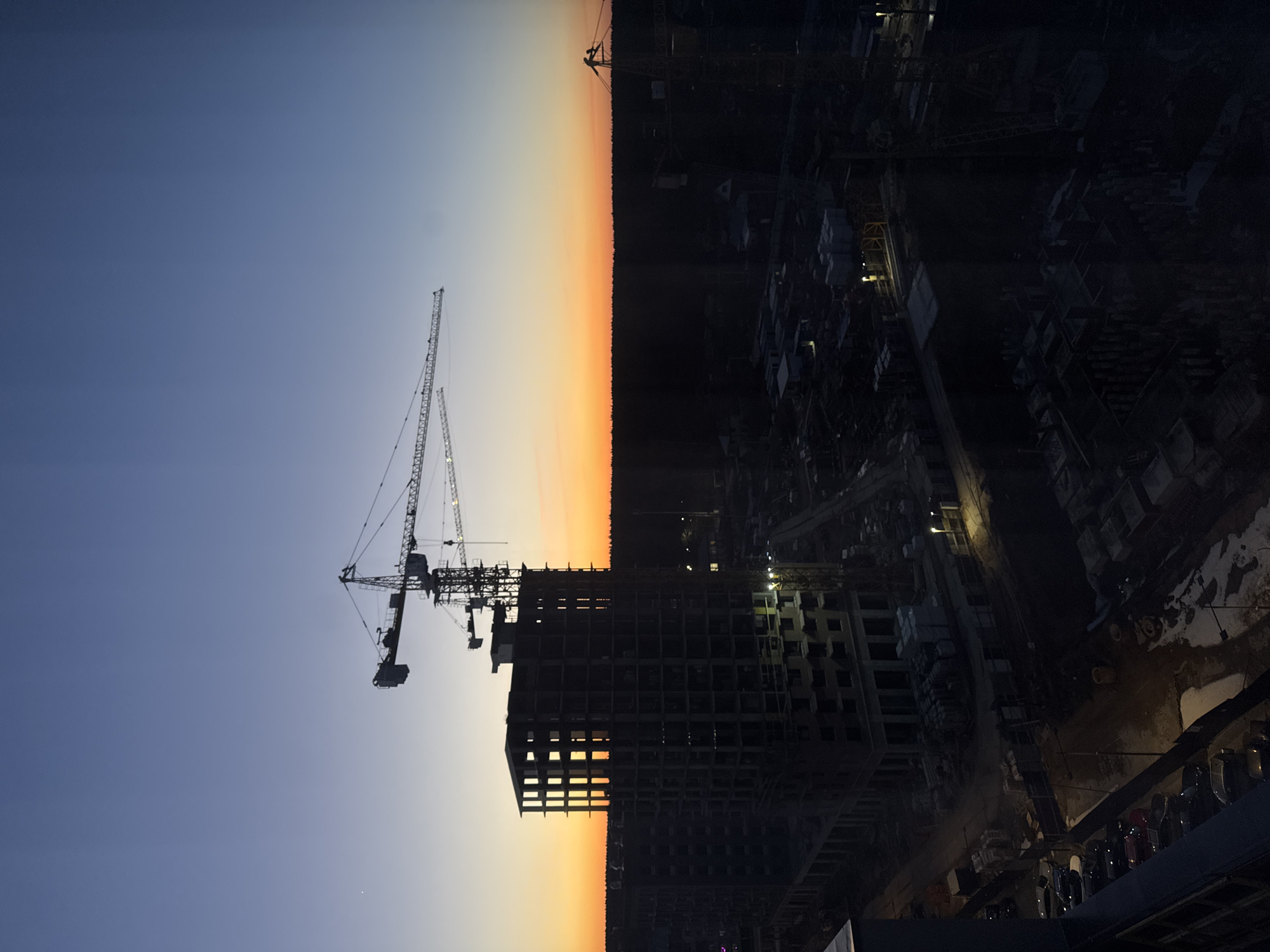 The image captures a construction site at dusk or dawn, potentially in Izhevsk, Russia. A towering construction crane stands prominently against a gradient sky, transitioning from blue to orange near the horizon, possibly indicating sunrise or sunset. The unfinished skeletal structure of a high-rise building is visible, with parts illuminated, hinting at ongoing work.
The right side of the image is predominantly dark, obscuring details of the surrounding environment, though construction materials and equipment can be seen. The overall setting suggests an urban construction project.