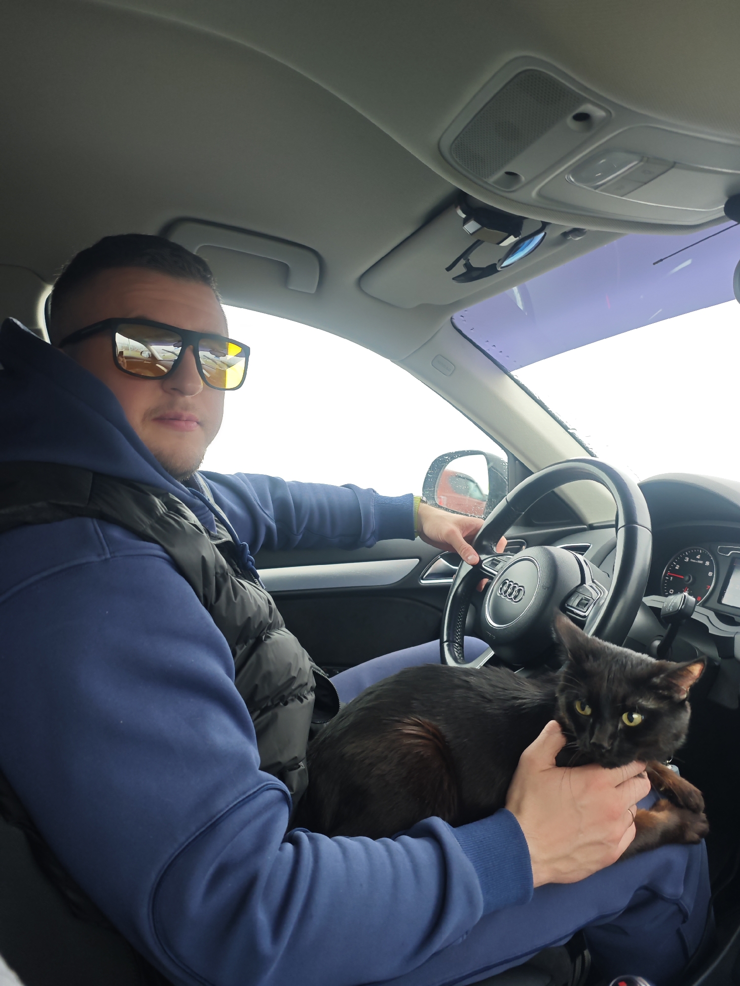 A man is seated in the driver's seat of a car, holding a dark-colored cat on his lap. He is wearing a navy blue hoodie, a black puffer vest, and sunglasses with yellow-tinted lenses, looking directly at the camera. His left hand rests on the steering wheel, which features a prominent "Audi" logo. His right hand gently cradles the black or very dark brown cat, which has bright yellow-green eyes and appears attentive, looking towards the camera as well.
The interior of the car is visible, including parts of the dashboard, speedometer (showing a low or zero speed), and the light-colored ceiling with overhead lights and a sunglass clip on the visor. The windshield shows a bright, overcast sky outside, suggesting daytime. A red car is faintly reflected in the side mirror. The scene depicts a casual moment between the man and his pet, possibly during a stop or a drive in Chaikovsky, Russia.