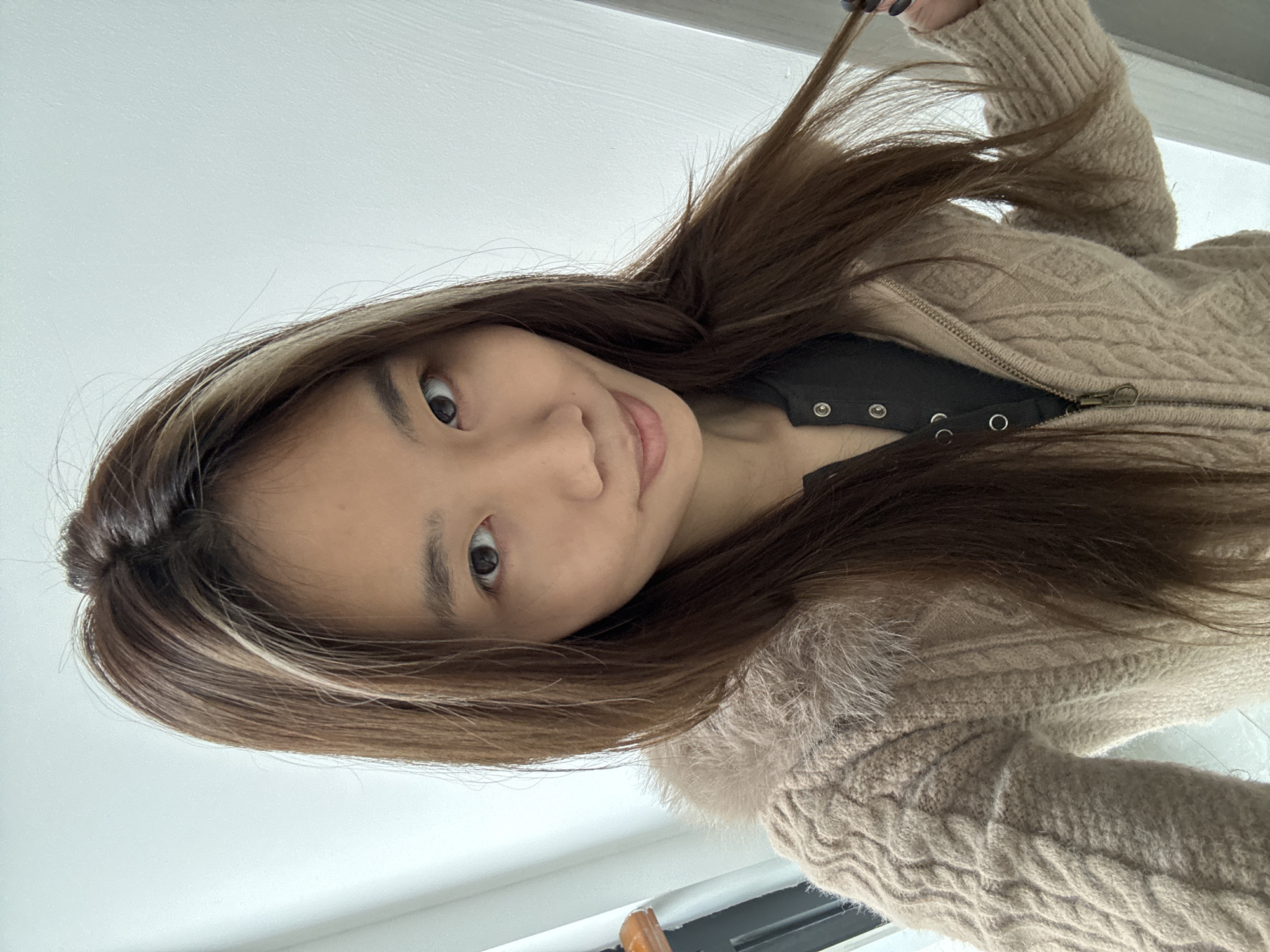 A medium close-up photograph features a young woman with long brown hair, slightly angled to the left. A strand of hair is held in her hand, obscuring part of the background. She is wearing a black top and a beige, cable-knit sweater with a fur-lined collar. The background is a plain white wall. No information indicates the location is in Nan'an District, China.