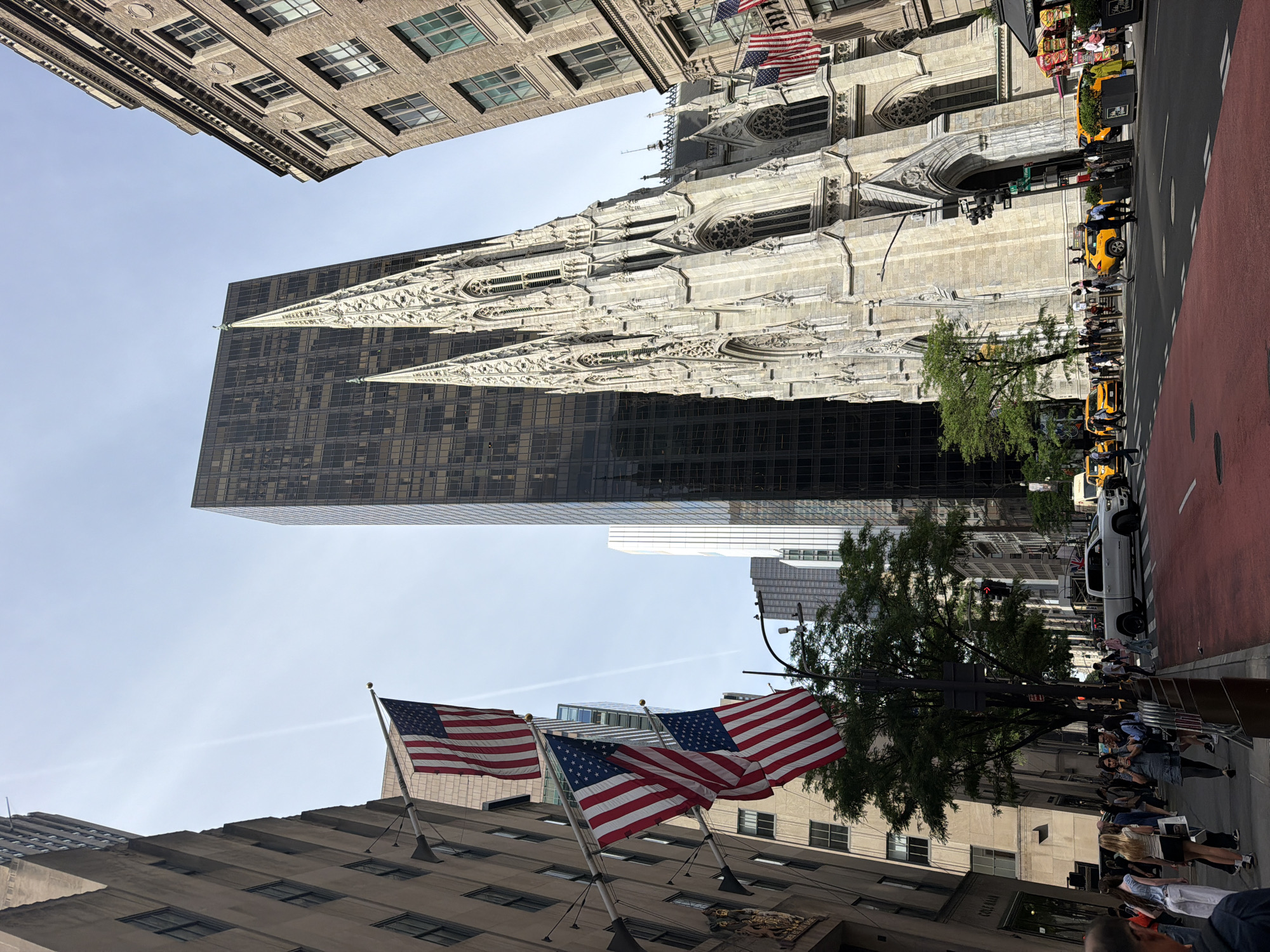 This image captures a vibrant street scene in New York City, dominated by a striking architectural contrast. In the mid-ground, the ornate, light-colored Gothic Revival architecture of St. Patrick's Cathedral rises grandly, featuring intricate spires, arches, and detailed stonework. Immediately behind it, a tall, dark, modern skyscraper with a gridded facade presents a stark contemporary counterpoint to the cathedral's historic design.
On the right side of the image, a busy street is visible with several iconic yellow taxi cabs, traffic lights, and numerous pedestrians strolling along the sidewalks. A distinct red-painted lane, likely a bike or bus lane, runs alongside the main road. Trees with green foliage are interspersed along the street, providing natural elements within the urban environment.
In the foreground and lower left, several American flags are prominently displayed on poles extending from a building. One flag, closer to the cathedral, is accompanied by a banner that partially reads "5TH AV" and "EST. 1953," strongly indicating the location as Fifth Avenue. Another building with multiple windows can be seen in the upper left, framing the clear, bright sky.
The scene depicts a sunny day in the city, with clear skies. People are visible engaged in daily routines, walking and interacting, creating a bustling atmosphere characteristic of a major metropolitan area. The overall impression is one of a dynamic urban landscape where history and modernity coexist.