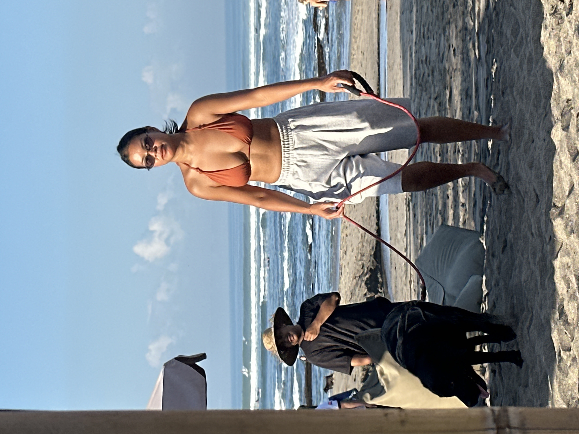 A woman in an orange bikini top and grey shorts walks a black dog on a red leash across a sandy beach. She wears sunglasses and her hair is pulled back. Beside her, slightly behind, a person in a black t-shirt and wide-brimmed hat walks the same dog. The beach has waves breaking in the background, and a large umbrella with a grey canopy is visible to the left. This scene appears to be in North Kuta, Indonesia.