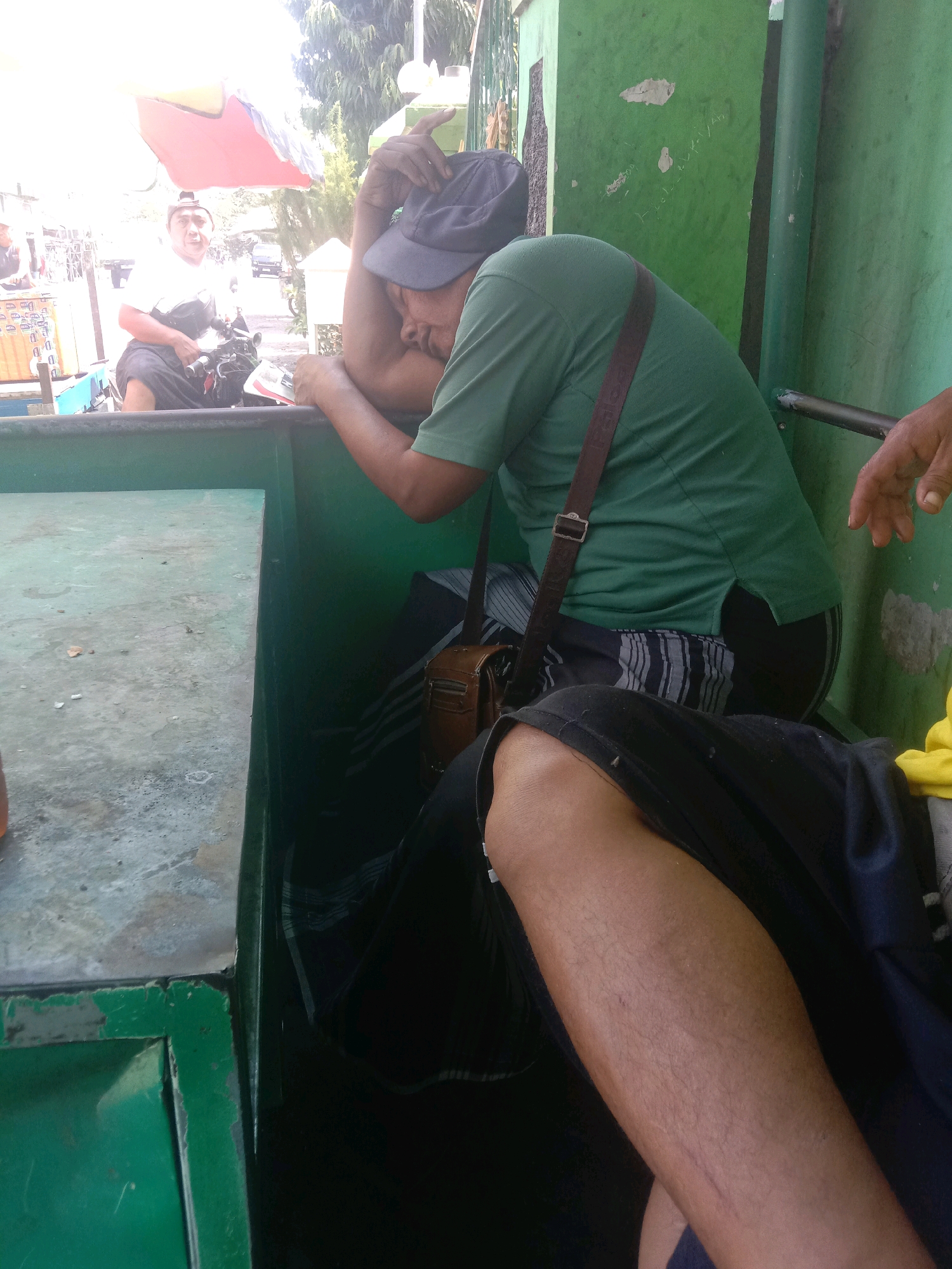In the city of Karang Anyar, Indonesia, a man wearing a blue cap and a green t-shirt is seen leaning against a green wall. He has a brown bag slung across his chest. To his left, another man in a white shirt and dark pants is seated on a motorcycle, with a red and yellow umbrella overhead. The foreground shows a green metal surface, possibly part of a vehicle or structure. The background features trees and a building. The scene appears to be outdoors, likely a street or market area, under daylight.