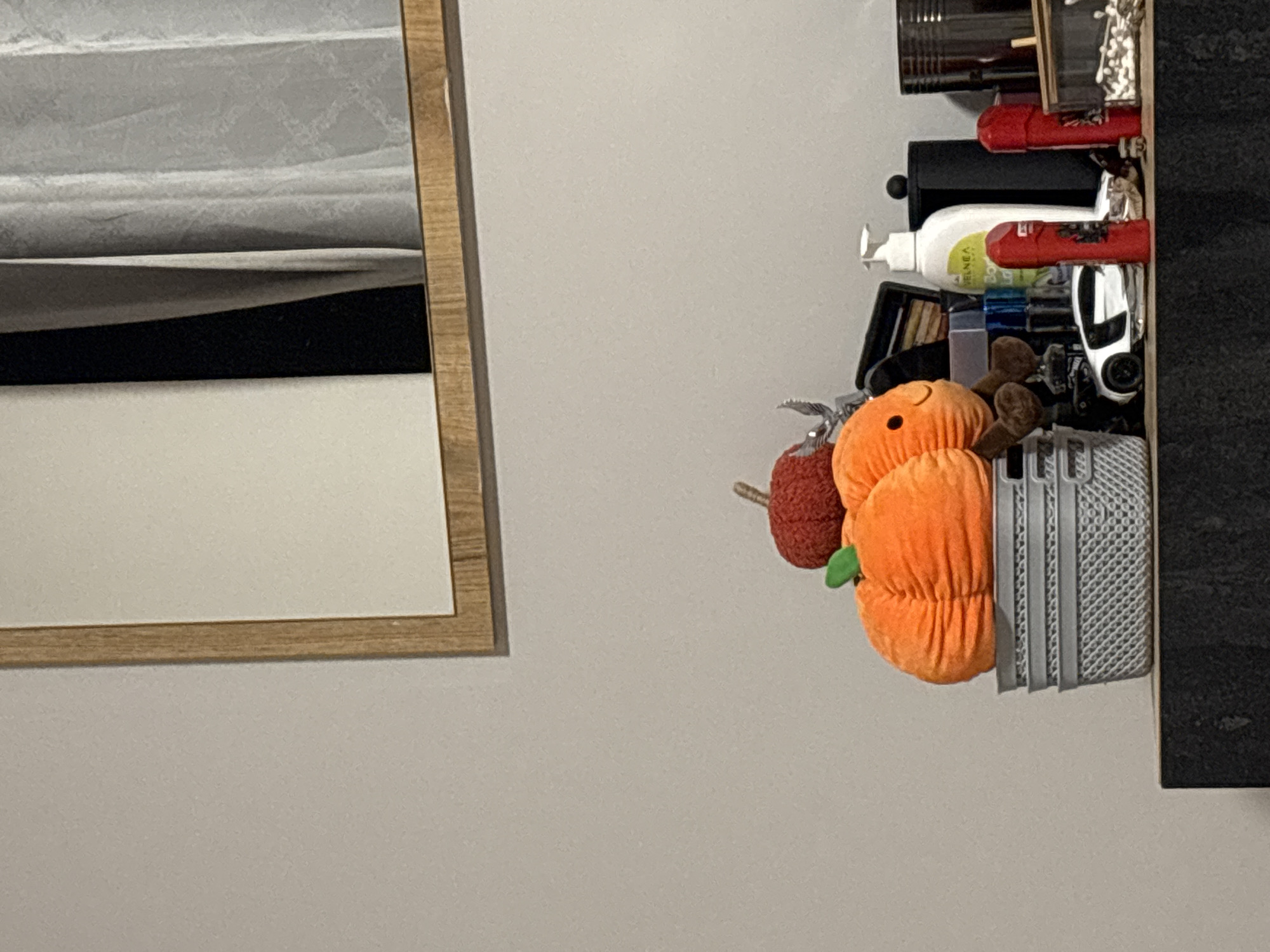 The image shows a shelf against a light gray wall. The shelf contains various items, including toiletries, small containers, a toy car, and plush toys shaped like an orange pumpkin and a red apple. A framed mirror is visible on the left side of the image. The location, Burgas, Bulgaria, cannot be determined from the provided image.
