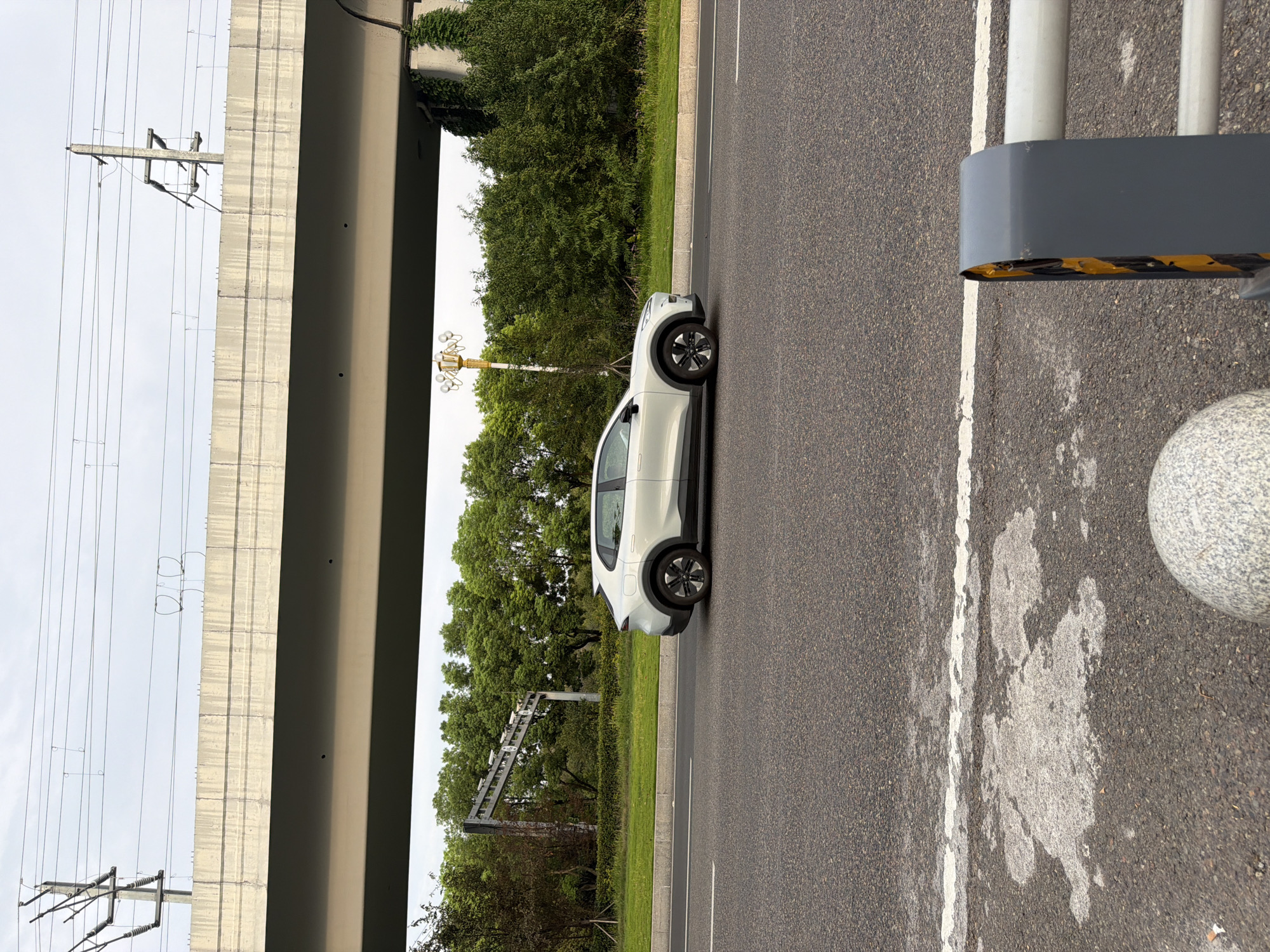 A silver electric crossover vehicle, identified as a Zeekr 001, is captured in profile, traveling northbound on an asphalt road. The vehicle is positioned in the right lane, with the road marked by a white dashed line on the left and a solid white line on the right. The surroundings depict a suburban or semi-urban environment, characterized by a clear sky overhead, with power lines and supporting structures visible above a concrete overpass. Lush green trees line the right side of the road, forming a dense backdrop. A decorative lamp post with multiple globes stands near the trees, suggesting a well-maintained roadside. In the foreground, a white painted section of the road is visible, along with the corner of a guardrail with yellow and black hazard markings. The image was taken in Jiangjin District, Chongqing, China.