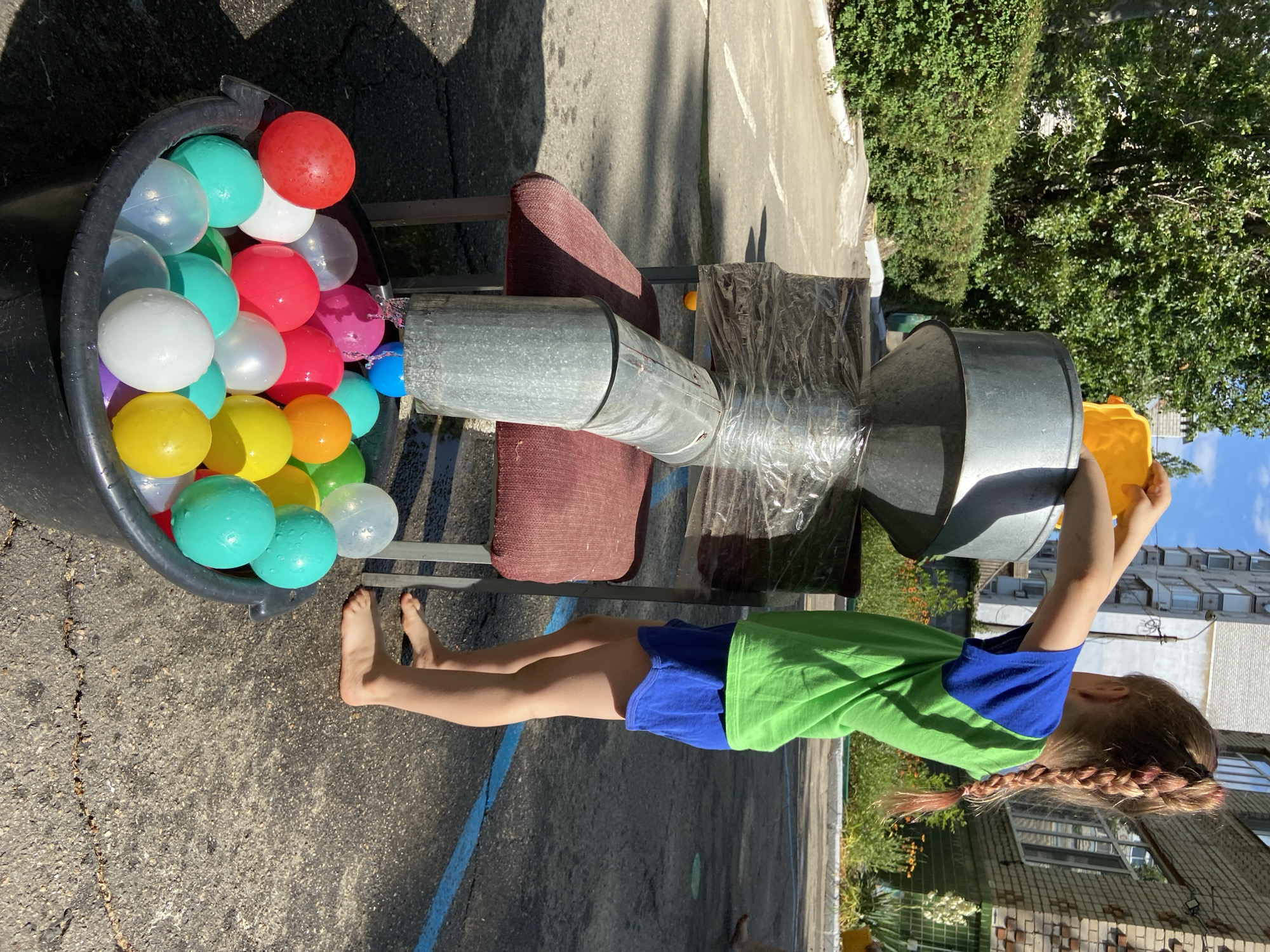In an outdoor urban setting, a young girl with a braided ponytail, wearing a green and blue shirt and blue shorts, stands barefoot on asphalt. She is holding a yellow plastic object and pouring its contents into a funnel made of galvanized metal, which is secured with plastic wrap to a chair. The funnel feeds into a metal pipe that is angled downwards. Below the pipe, a black plastic bucket is filled with a variety of colorful balls, including red, white, pink, purple, green, teal, and yellow. The background shows trees, a hedge, a brick building, and a multi-story apartment building under a clear blue sky. The scene appears to be part of an outdoor activity or game.